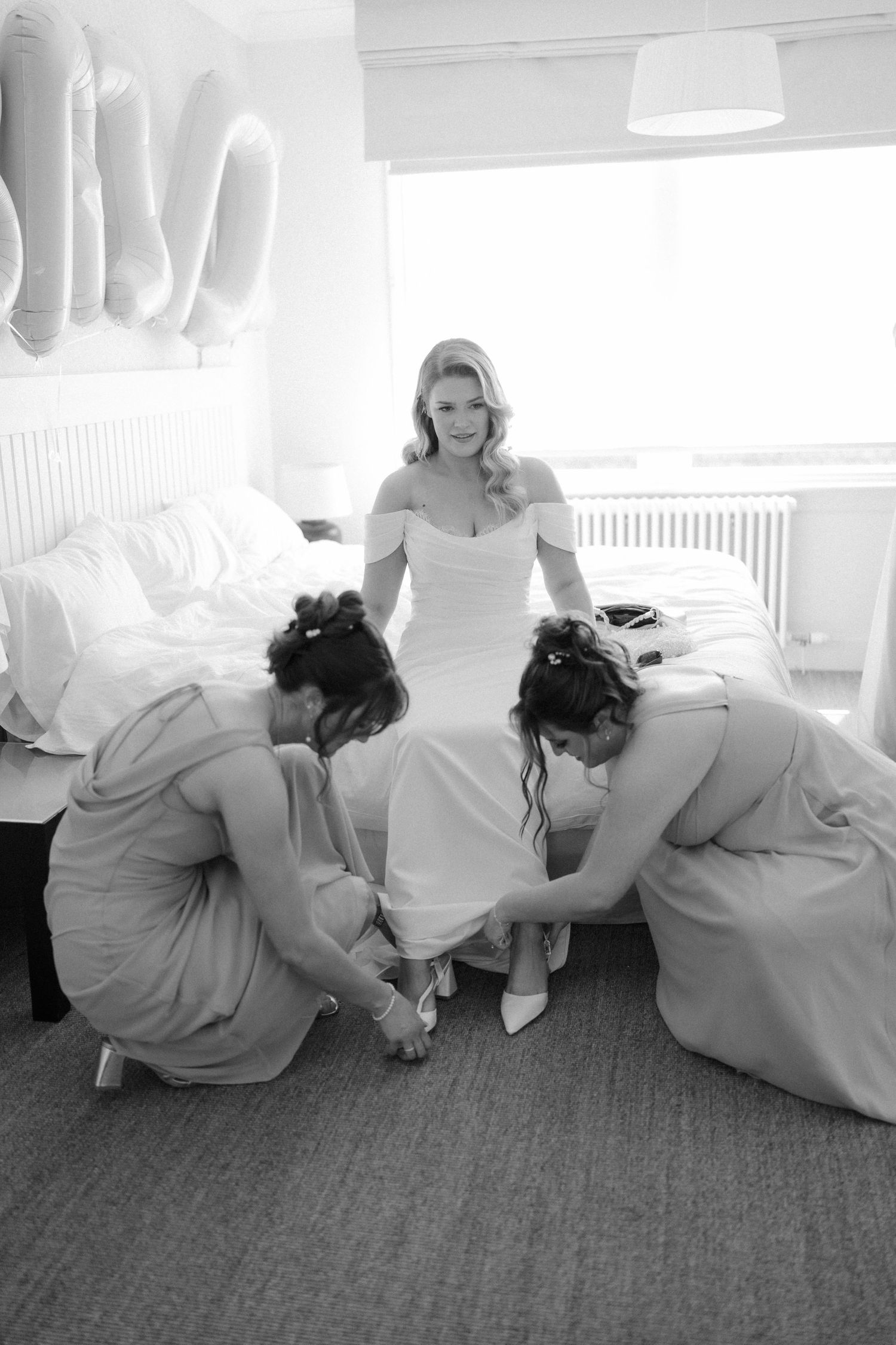 Bride in white gown sits on bed while two bridesmaids help put on her shoes before wedding.