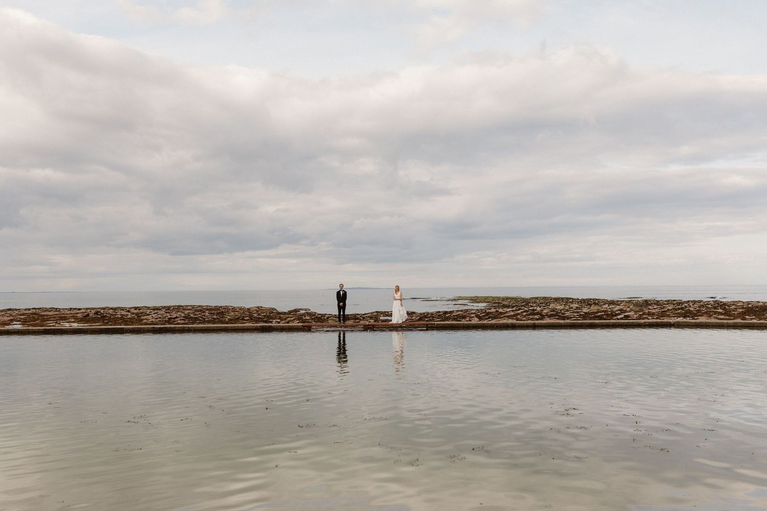 Bride and groom standing on coastal stone pier with dramatic cloudy sky and calm water reflection.