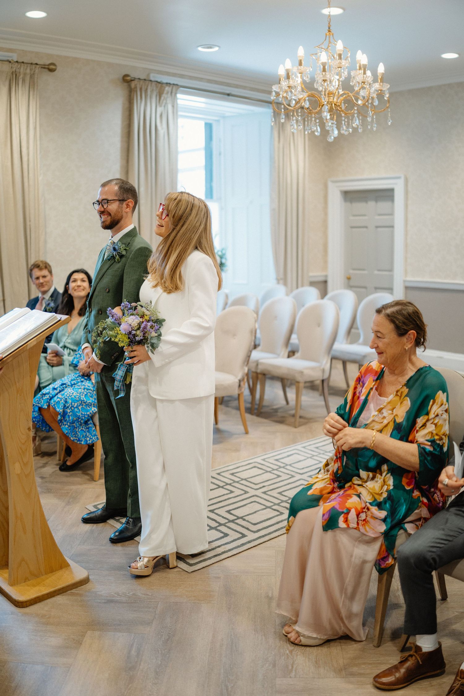 Elegant bridal suite with chandelier and white chairs shows people seated in conversation before a wedding ceremony.
