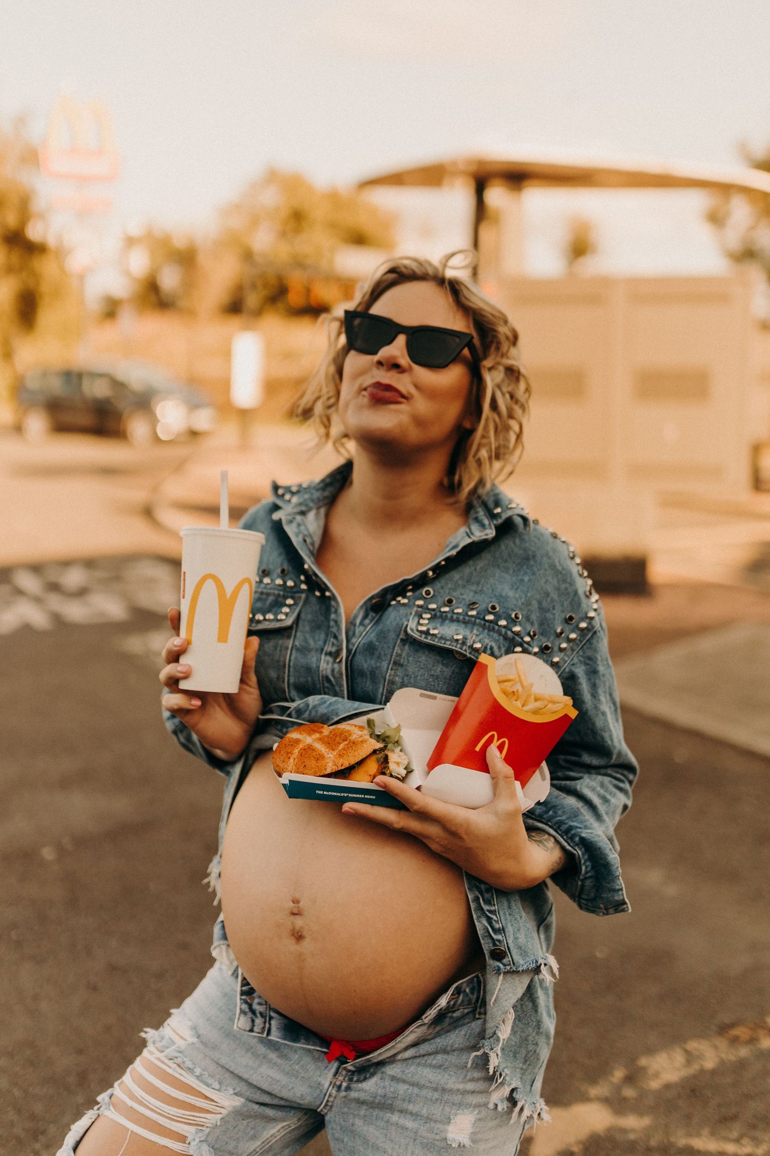 Maternity photoshoot featuring McDonald's food and denim outfit in warm sunset lighting.