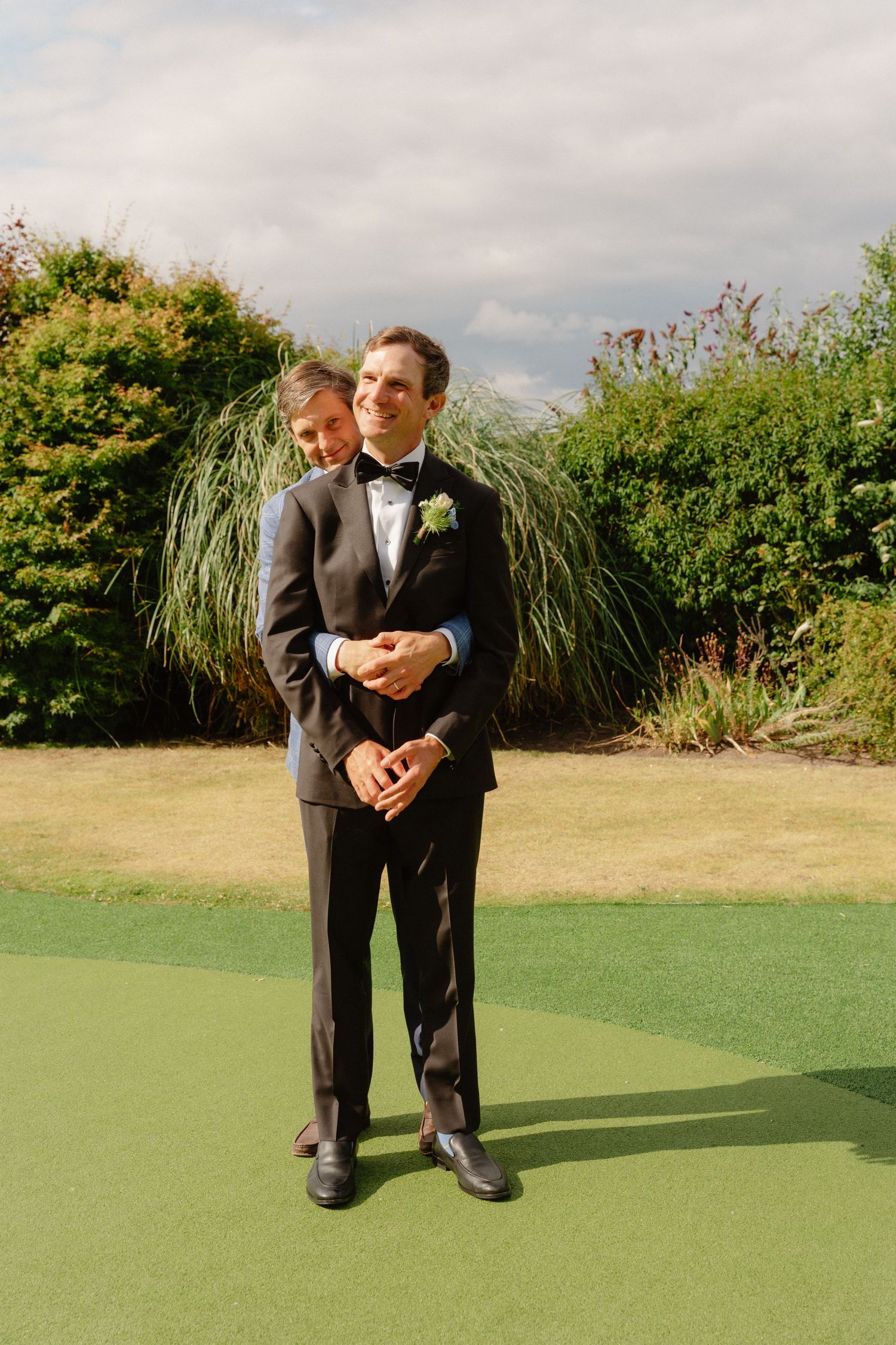 Groom in black tuxedo with boutonniere stands smiling on green lawn with lush garden background.