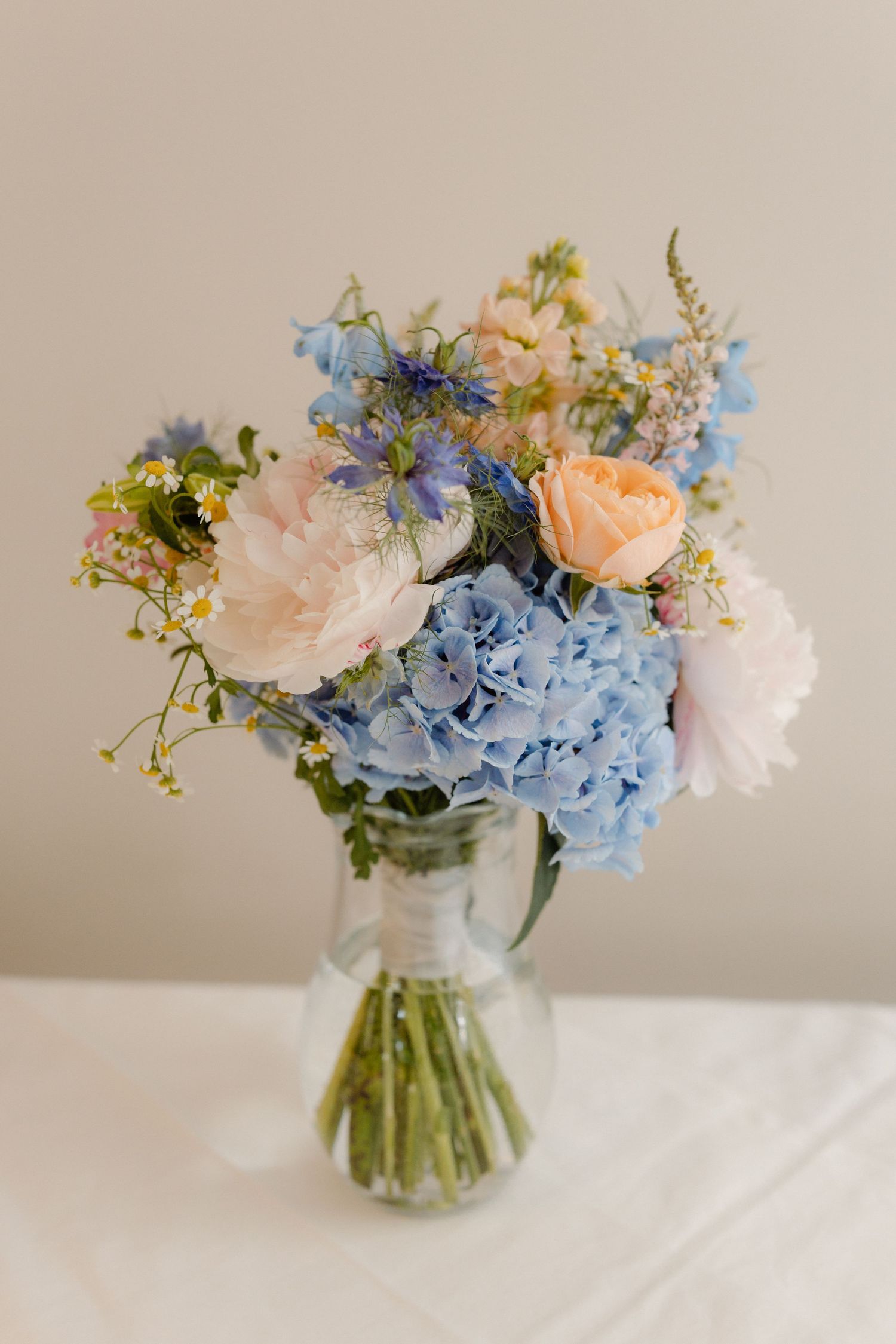 Beautiful bridal bouquet featuring blue hydrangeas, peach roses, and wildflowers in a glass vase.