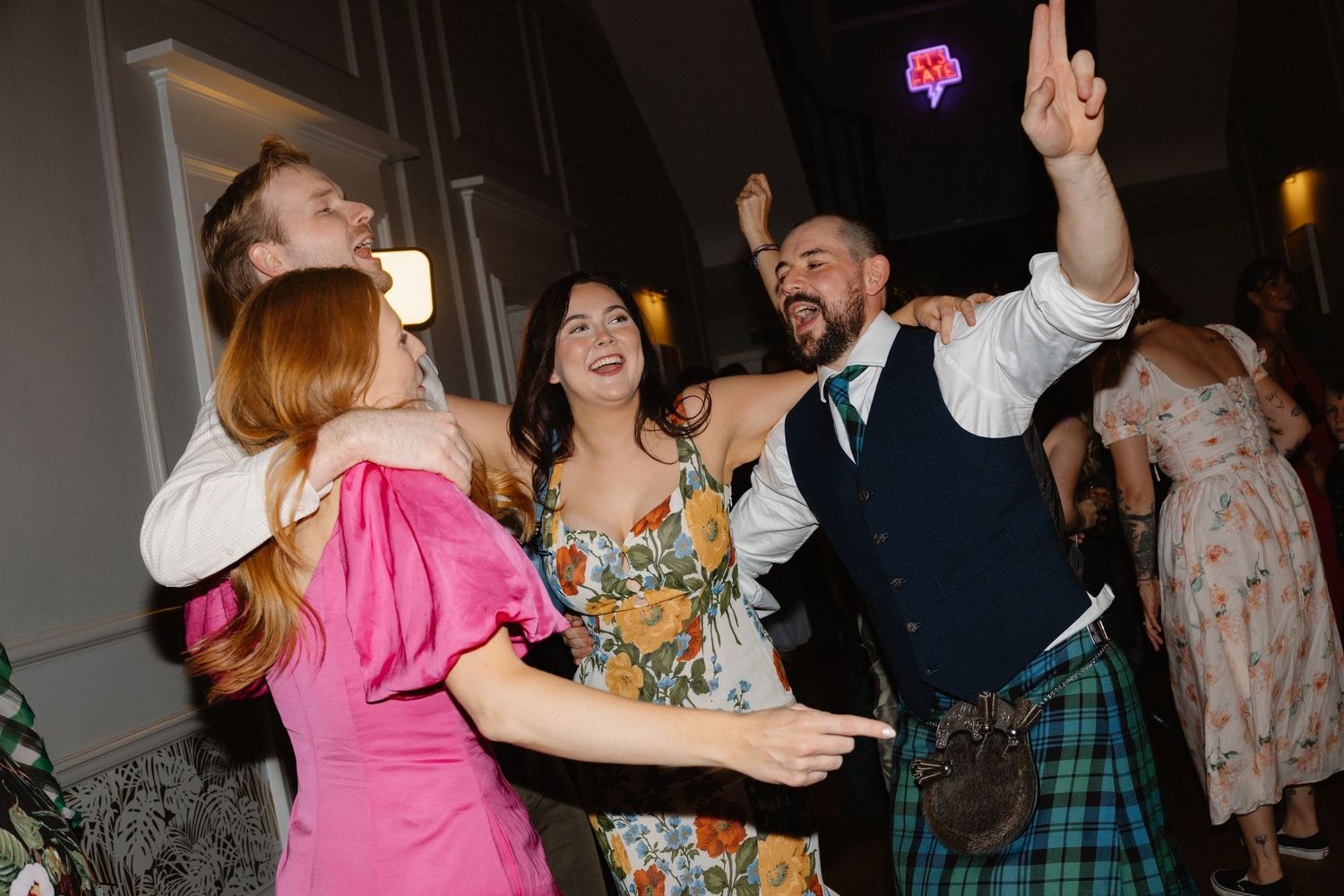 Guests in kilts and colorful dresses celebrate joyfully on the dance floor at a Scottish wedding reception.