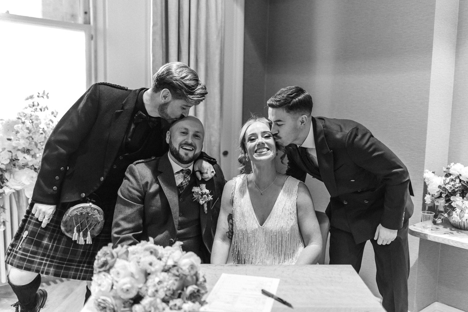 Black and white wedding photo of bride and groom laughing with two guests kissing their cheeks at signing table.