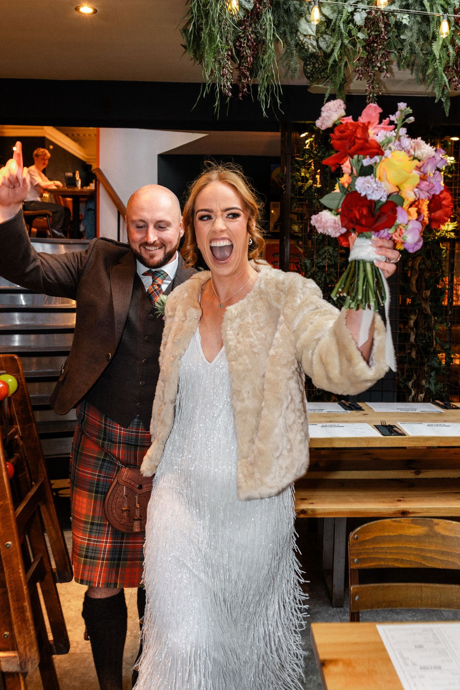 Excited bride in fringed dress and fur jacket raising colorful bouquet with groom in kilt.