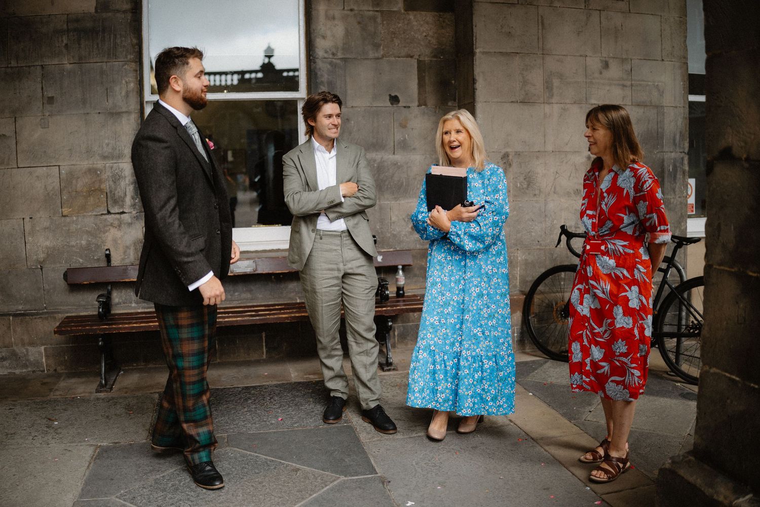 Wedding party in formal attire chatting outside stone building, woman in blue dress holding tablet.