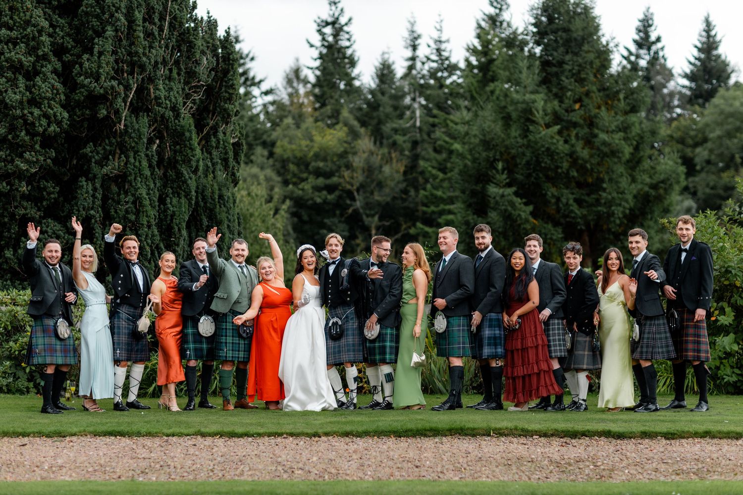 Large wedding party group photo on lawn, guests wearing Scottish kilts and colorful formal attire with evergreen trees behind.