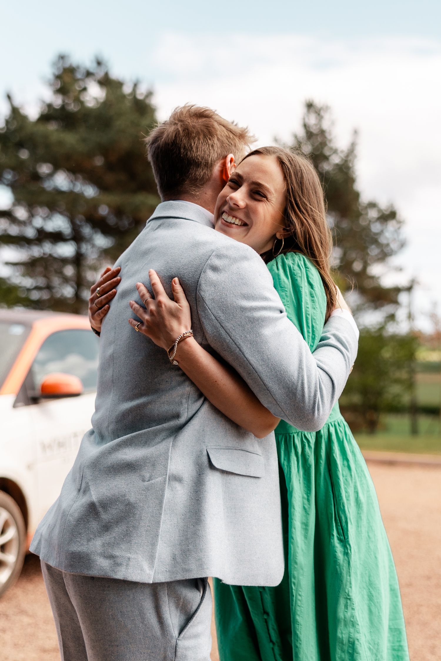 Smiling woman in green dress hugging man in grey suit outdoors near vintage orange car.