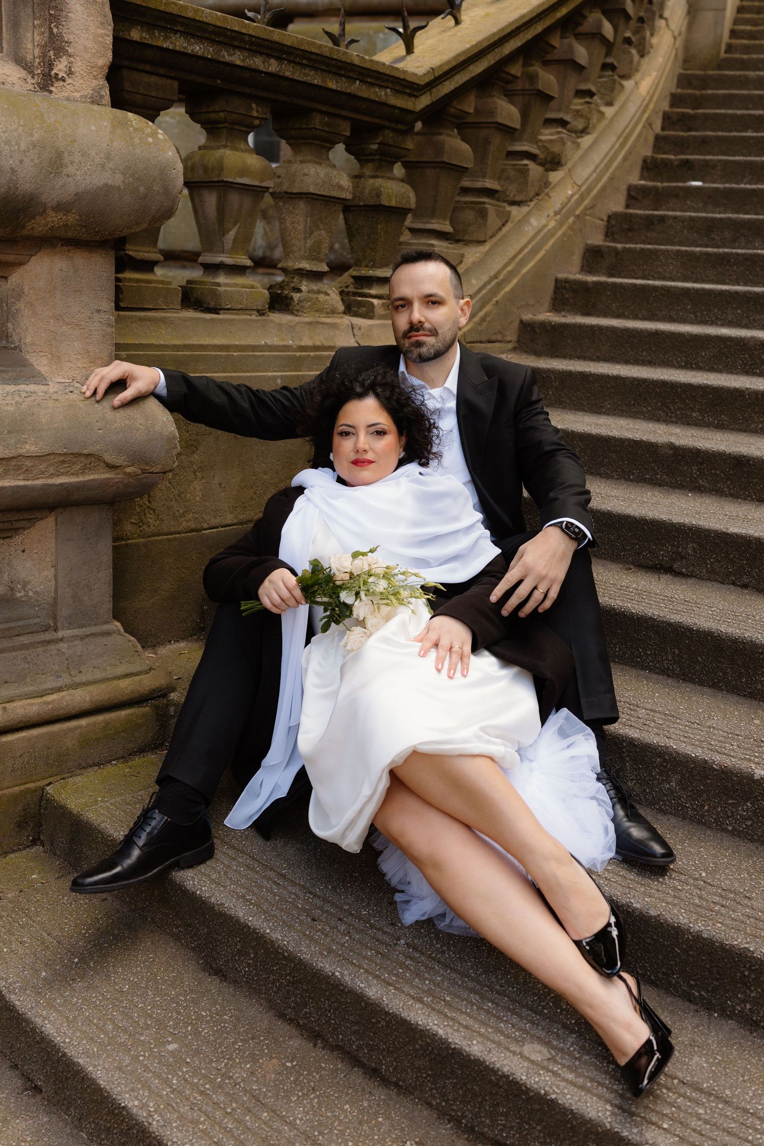 Bride and groom sitting on stone steps of a grand staircase, bride holding white roses bouquet.