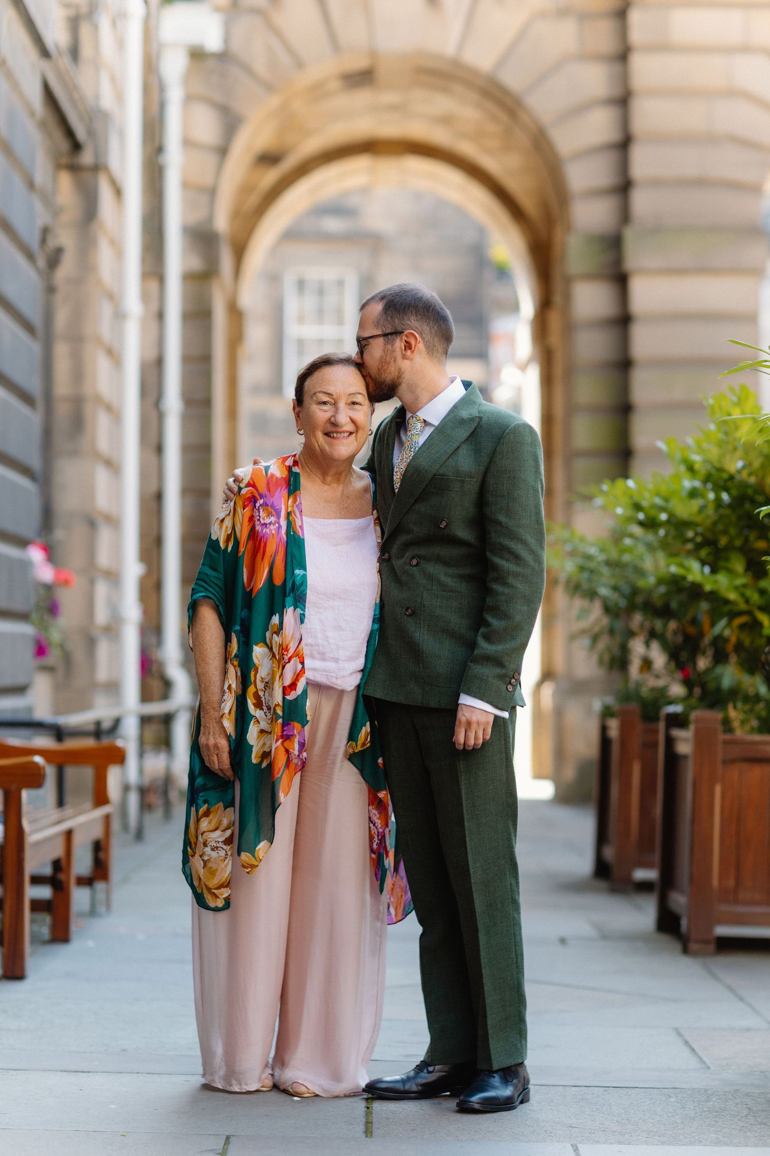 Man in green suit kissing older woman in floral kimono outdoors under stone archway.