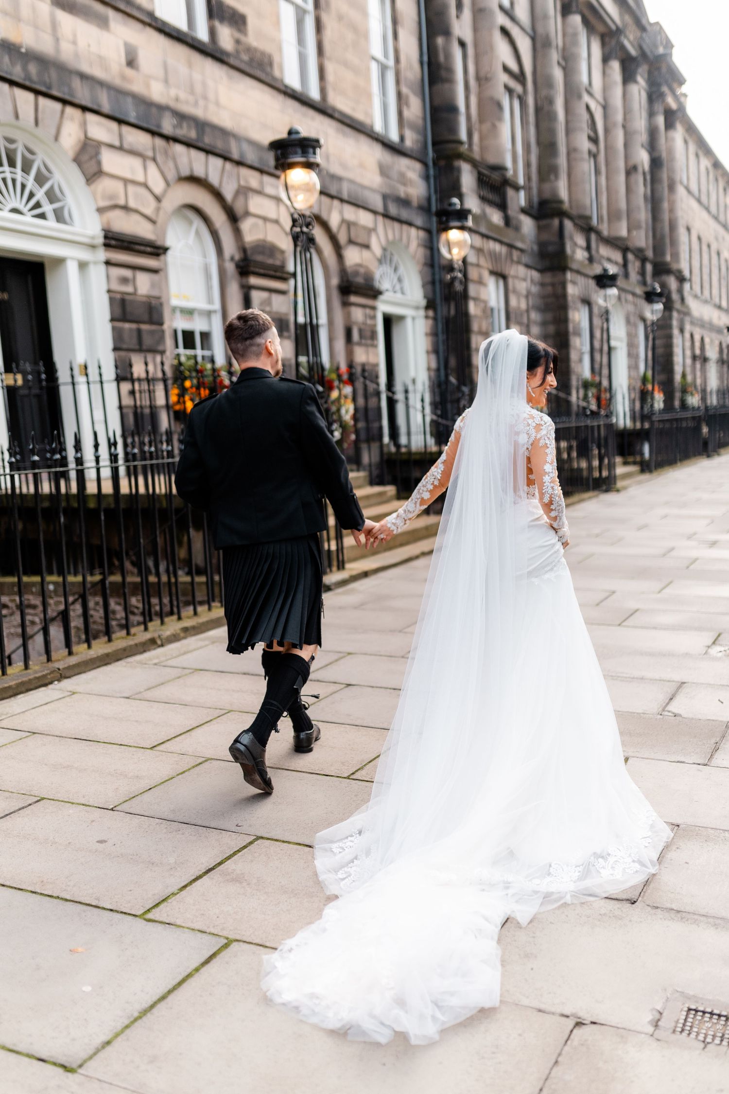 Bride in lace gown and groom in kilt walking down historic Edinburgh street.