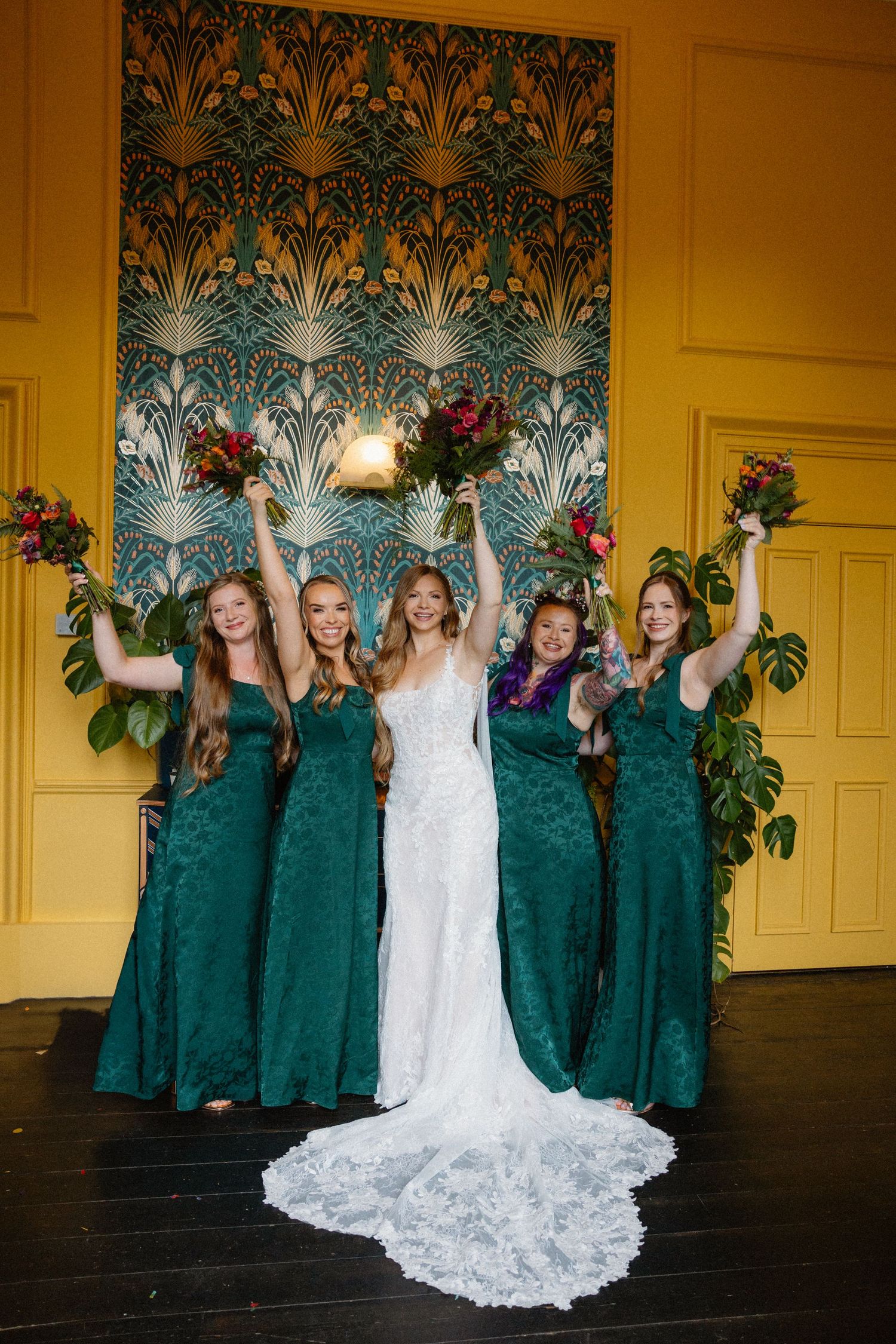 Bridesmaids in emerald green dresses posing with bride in white gown against patterned wallpaper.
