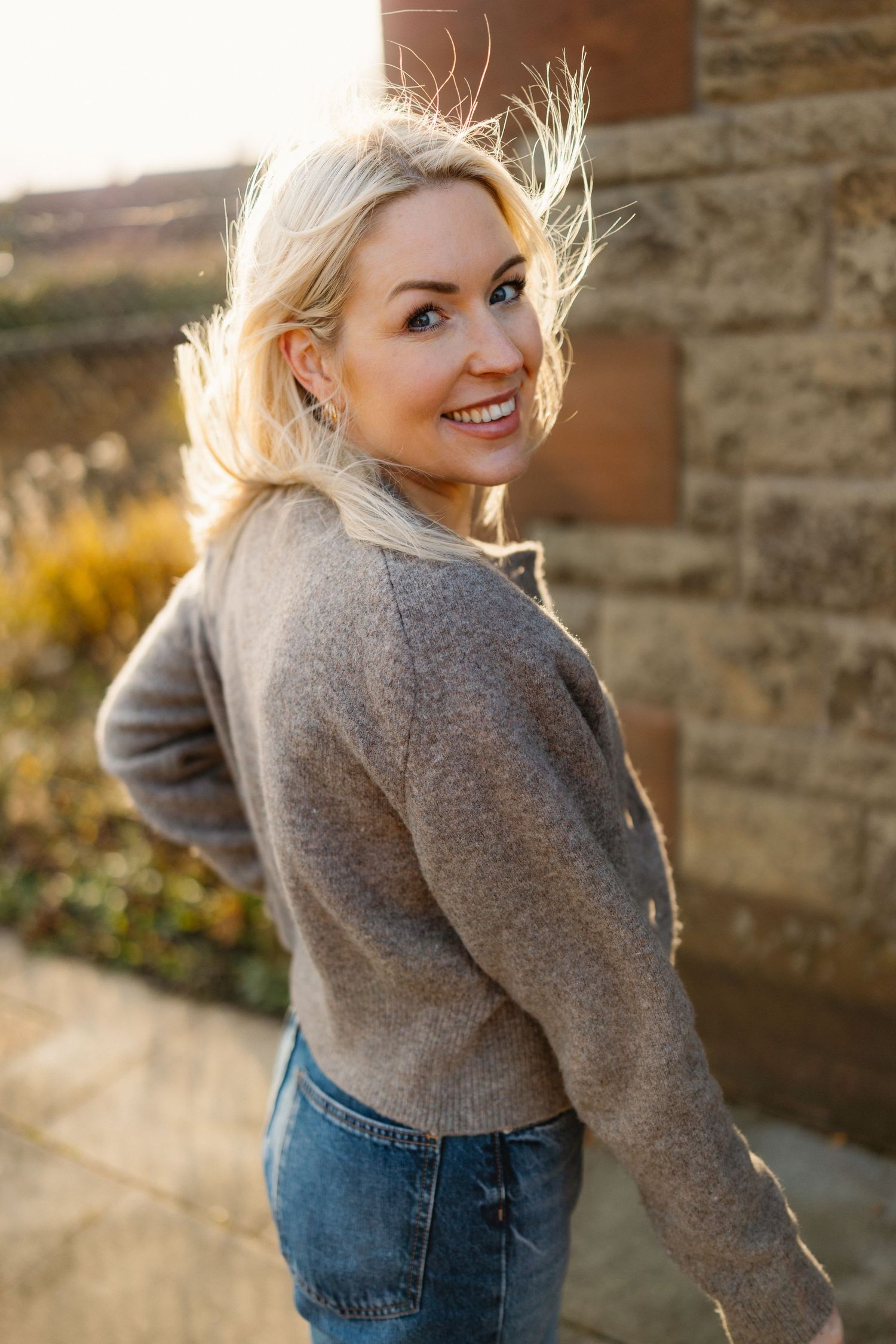 Person wearing grey sweater and jeans poses playfully against brick wall in natural lighting.