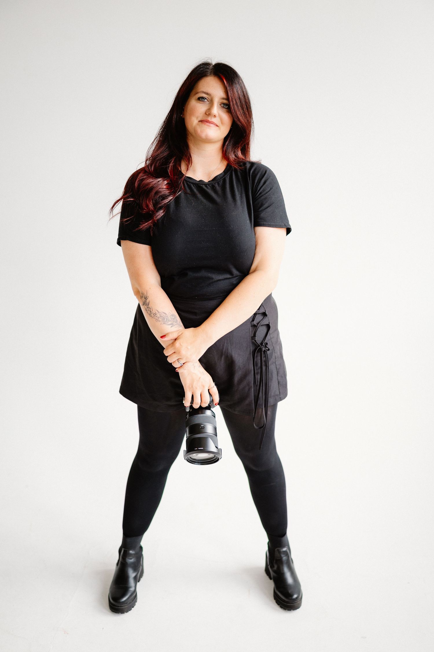 Full-length portrait of person in all black outfit holding shoes against white background.
