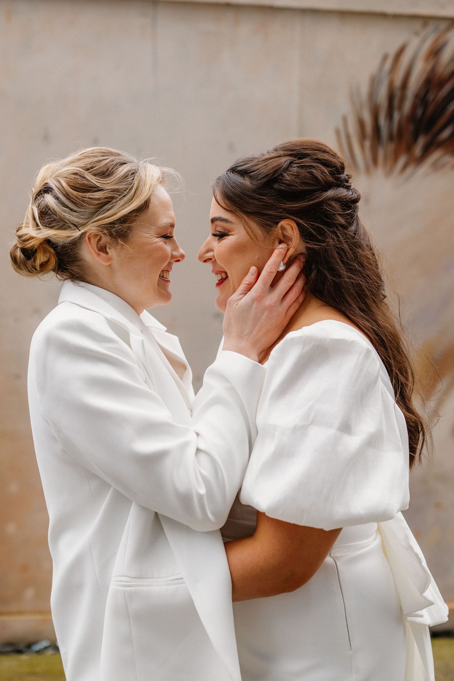 Two brides in white outfits share a joyful moment, smiling and embracing on their wedding day.