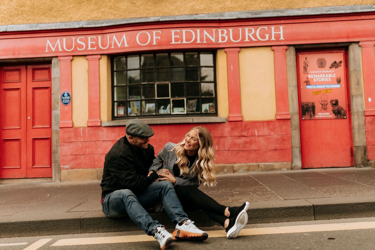 Two people sitting outside the Museum of Edinburgh building with its distinctive red facade.