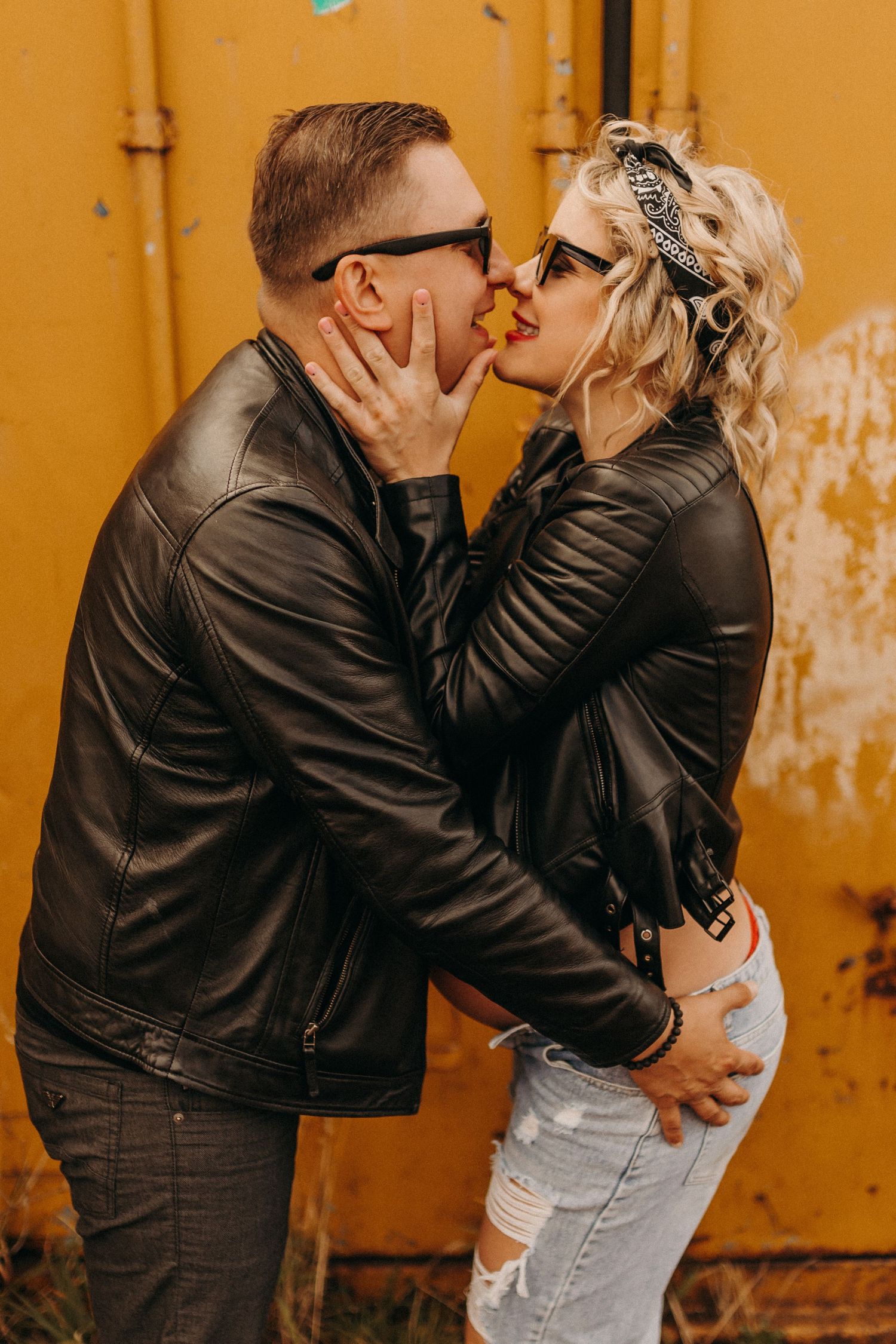 Romantic couple in leather jackets embracing against vibrant orange wall.