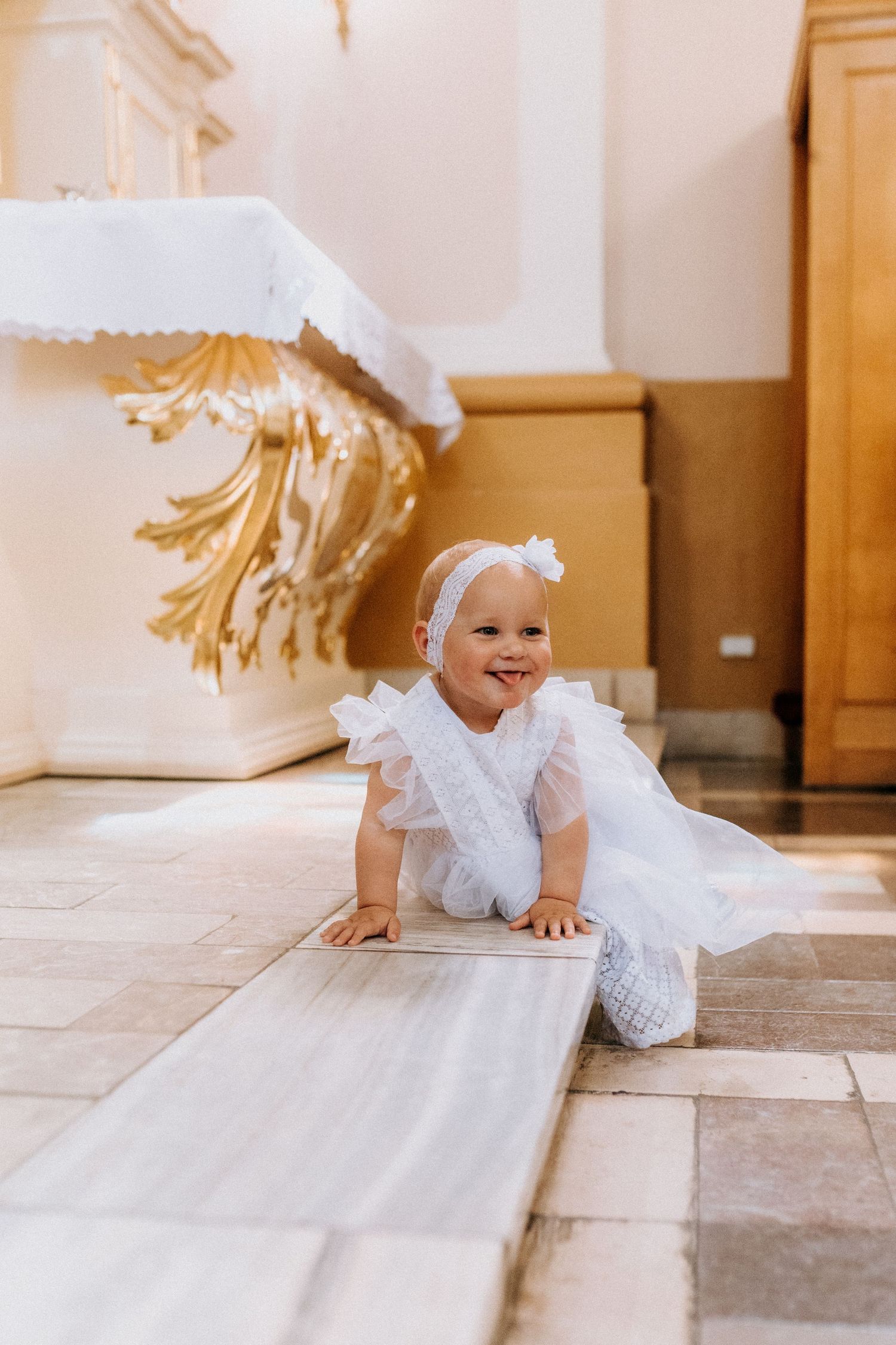 Baby in white dress crawling on tiled floor during special occasion.