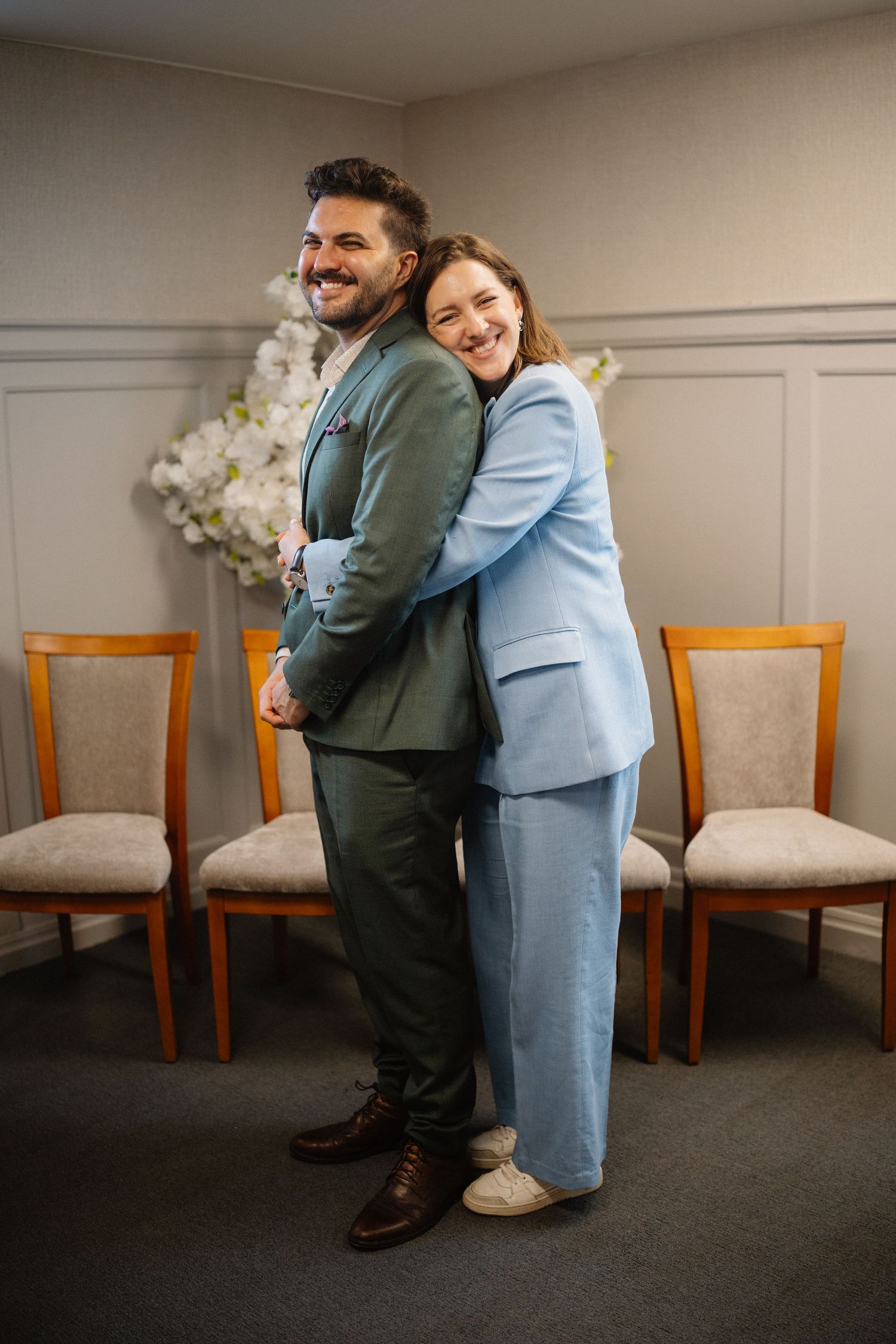 A couple in coordinating suits stands back to back in an elegant room with wooden chairs and wainscoting.