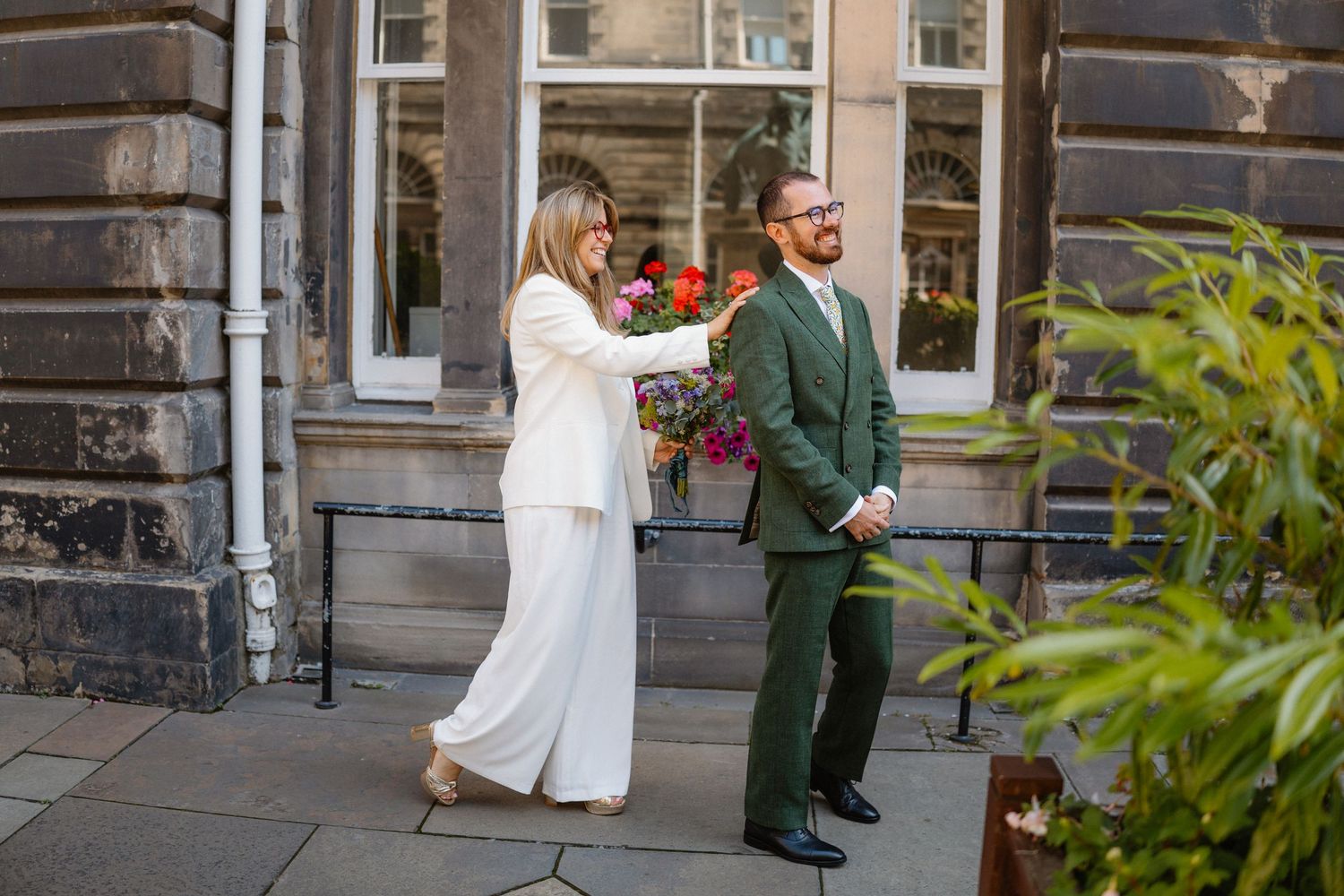 A couple in contrasting white and green formal attire share a joyful moment outside a historic stone building.