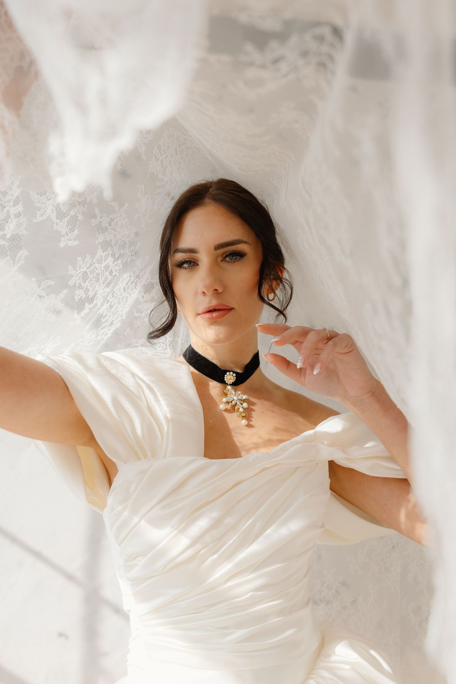 Elegant bride in white satin gown with black velvet choker necklace, surrounded by flowing lace fabric in bright light.