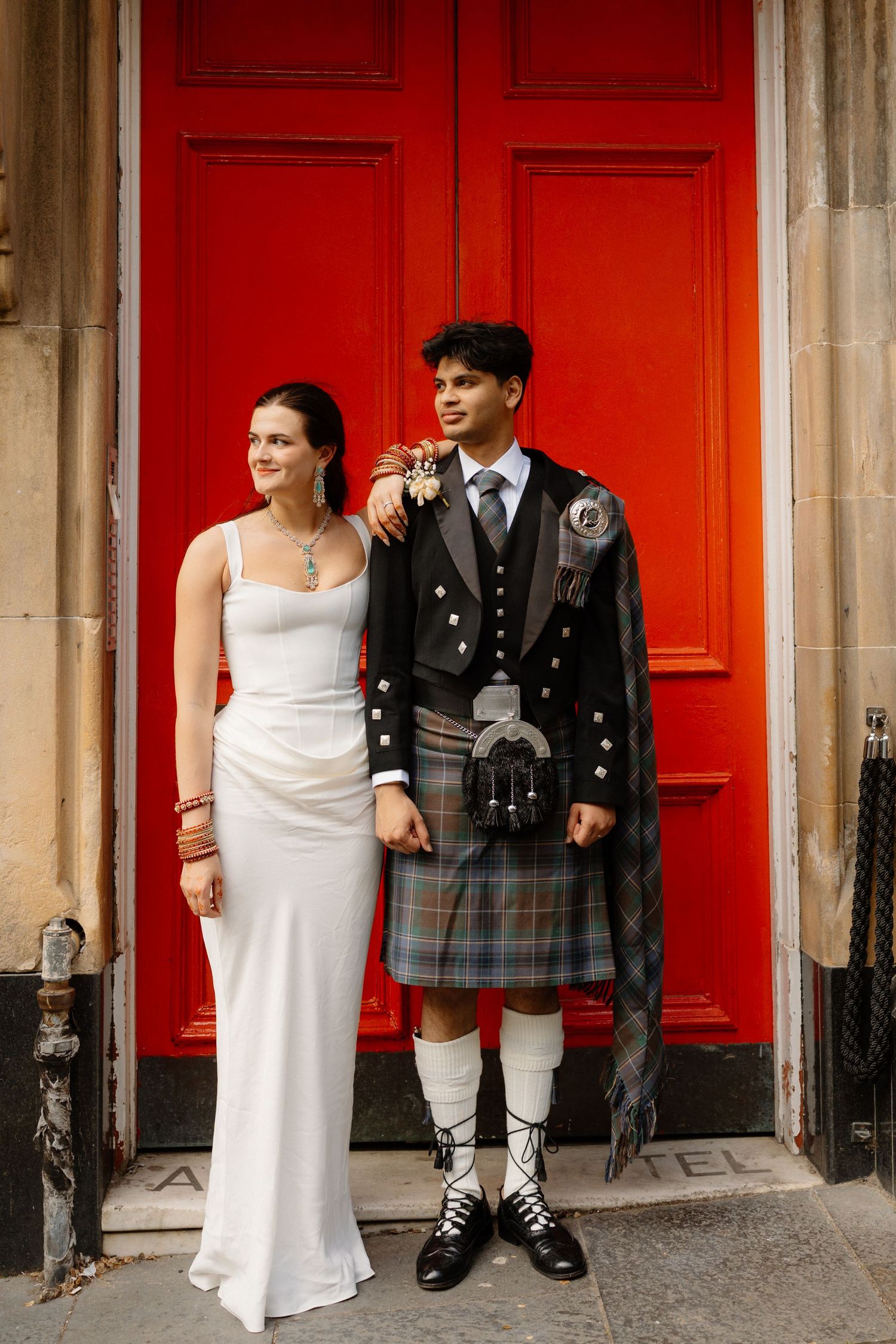 Bride in white gown and groom in traditional Scottish kilt standing before a vibrant red door.