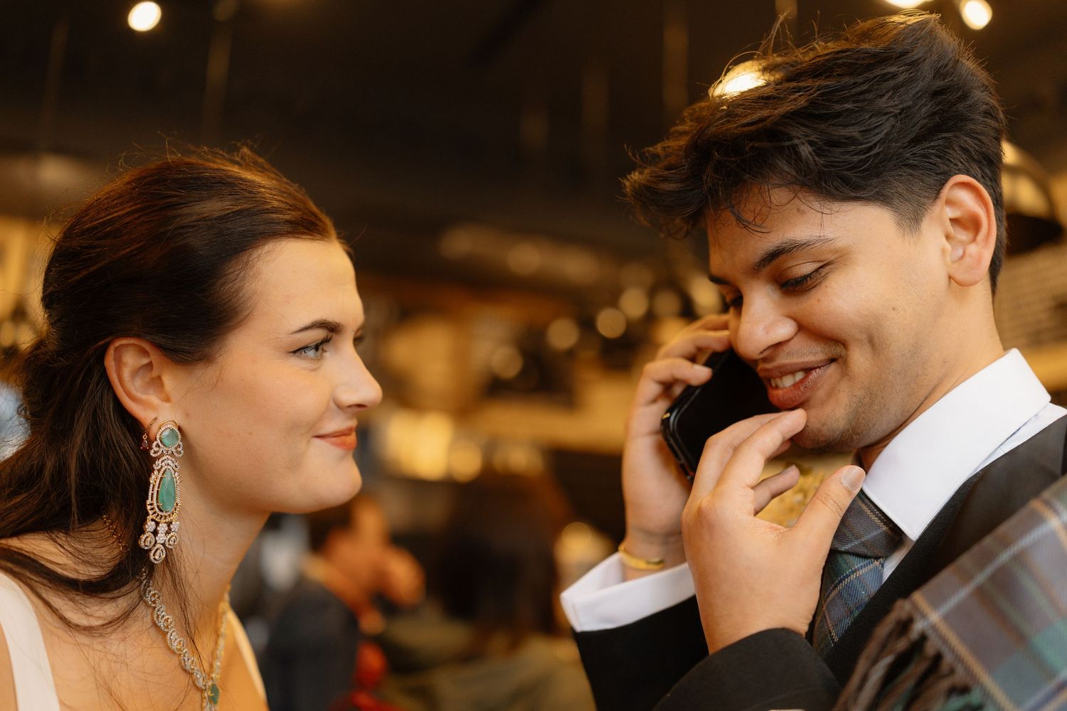 Elegant couple at a formal event, woman in jewelry smiling at man in suit talking on phone.