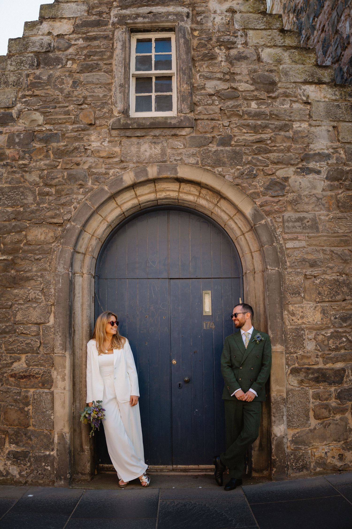 Bride and groom posing in front of a rustic stone archway with a blue wooden door on their wedding day.