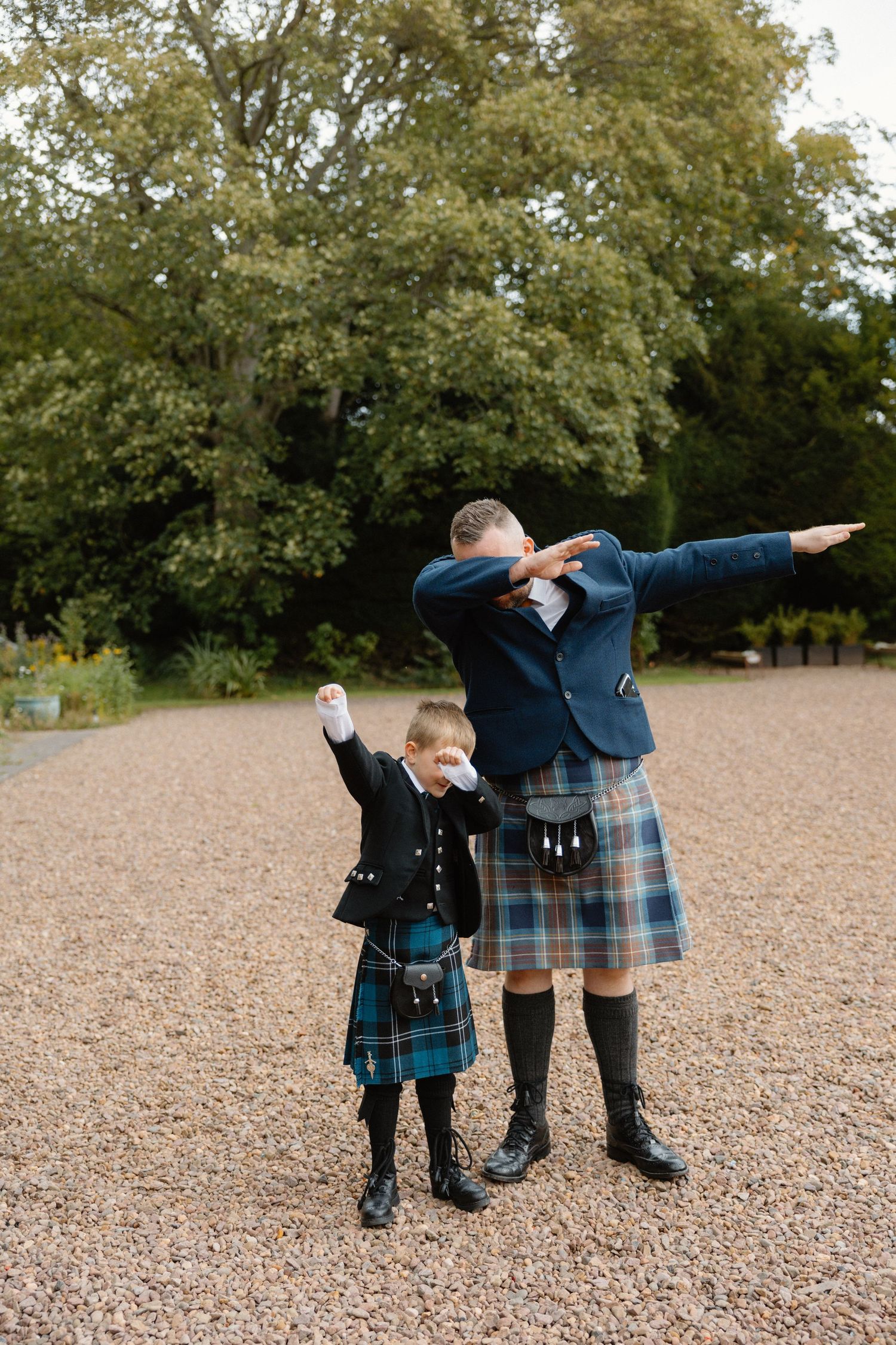 Man and boy in matching Scottish kilts doing a dab pose outdoors on gravel path.