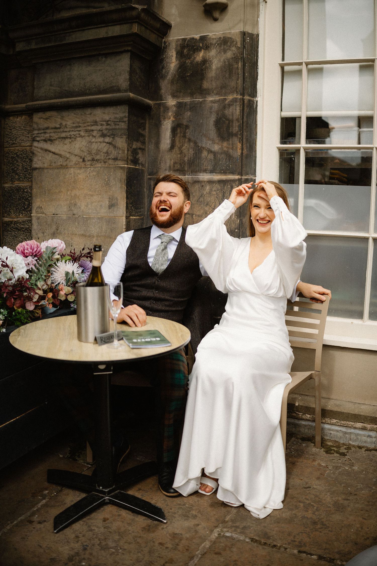 Laughing bride and groom sitting at outdoor table with champagne bucket at Scottish wedding venue.