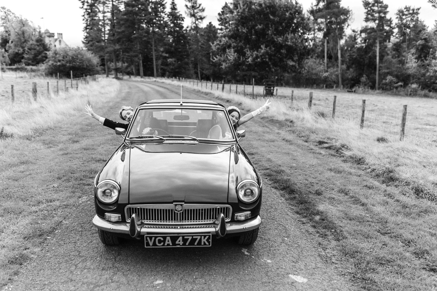 Black and white photo of couple posing in a classic MG convertible on a rural tree-lined road.