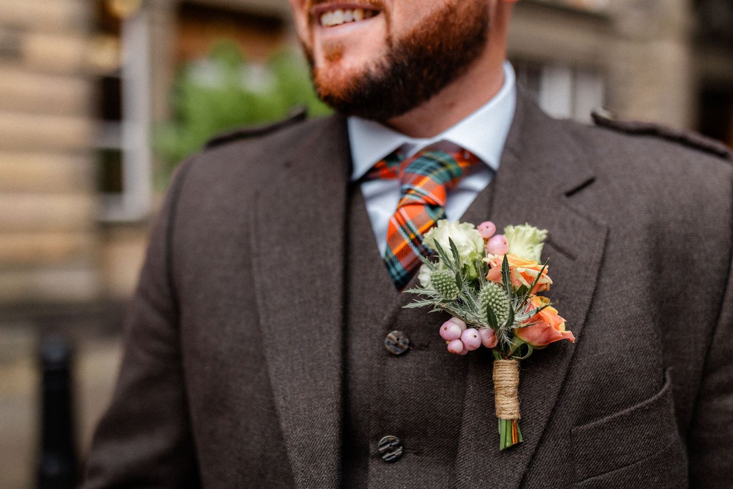 Smiling groom in brown tweed suit with tartan tie and colorful floral boutonniere on wedding day.