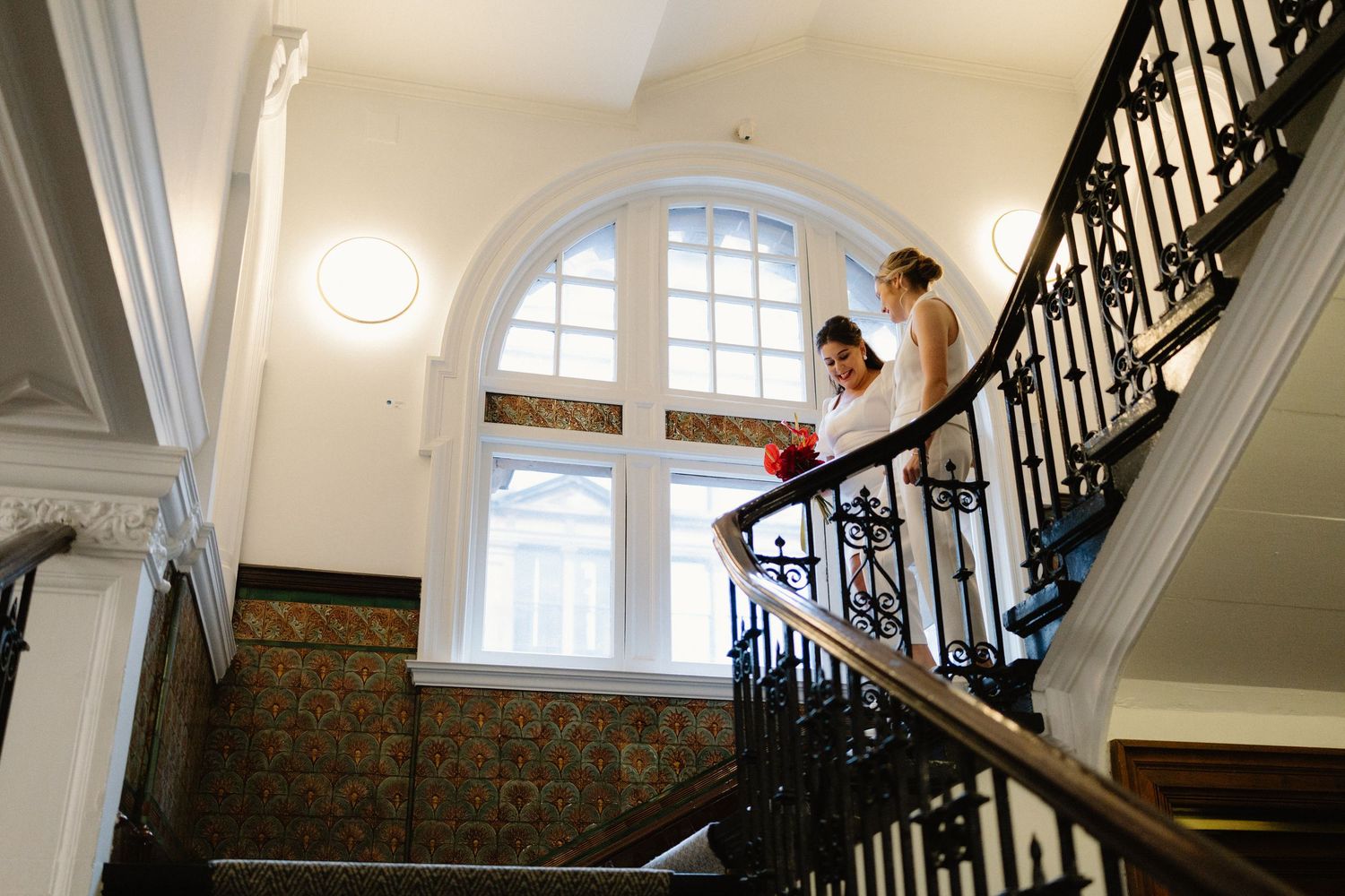 Two brides in white descend an elegant staircase with ornate iron railings and arched window in a historic venue.