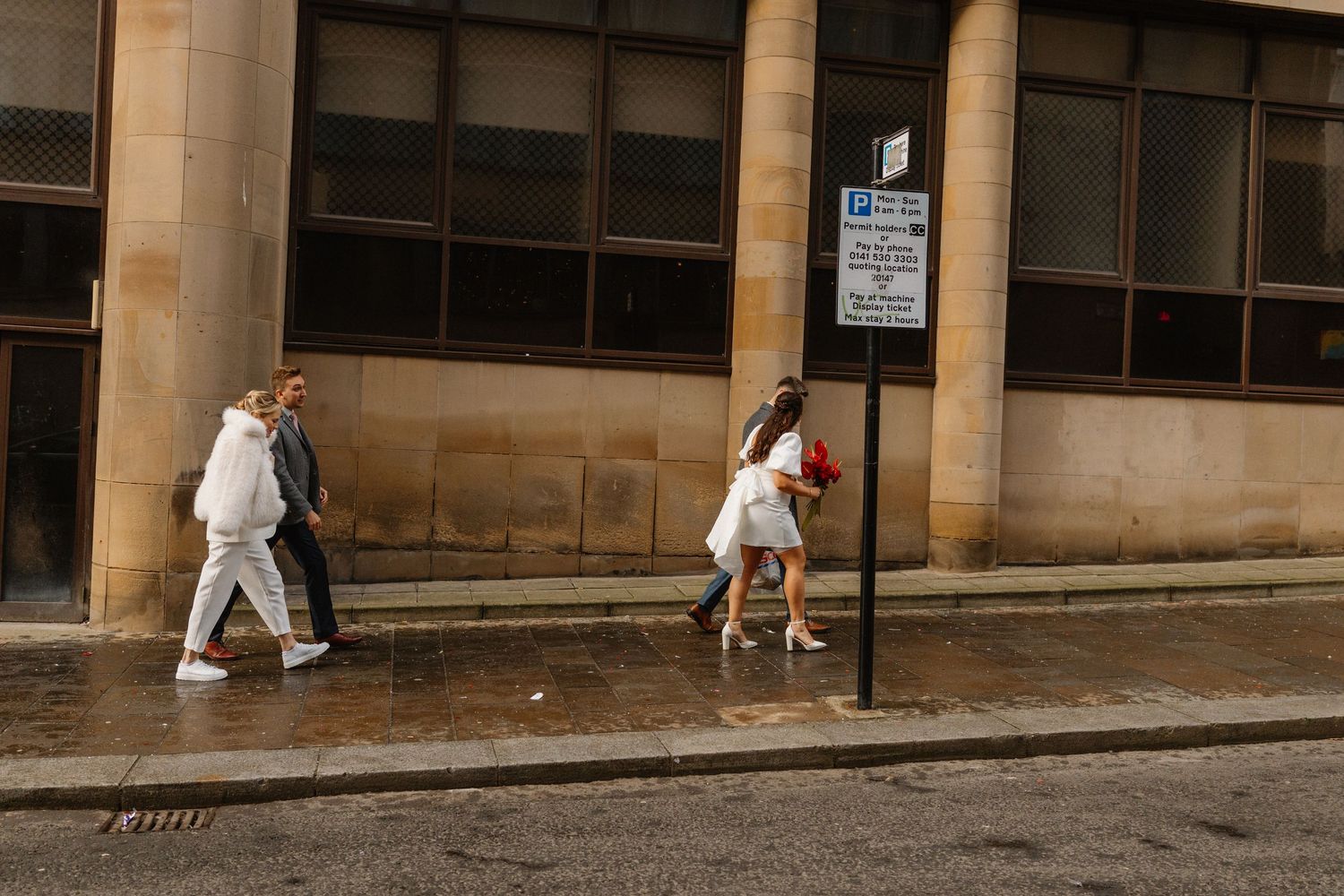 Three people walking on a wet urban sidewalk past a stone building with large windows and a parking sign.