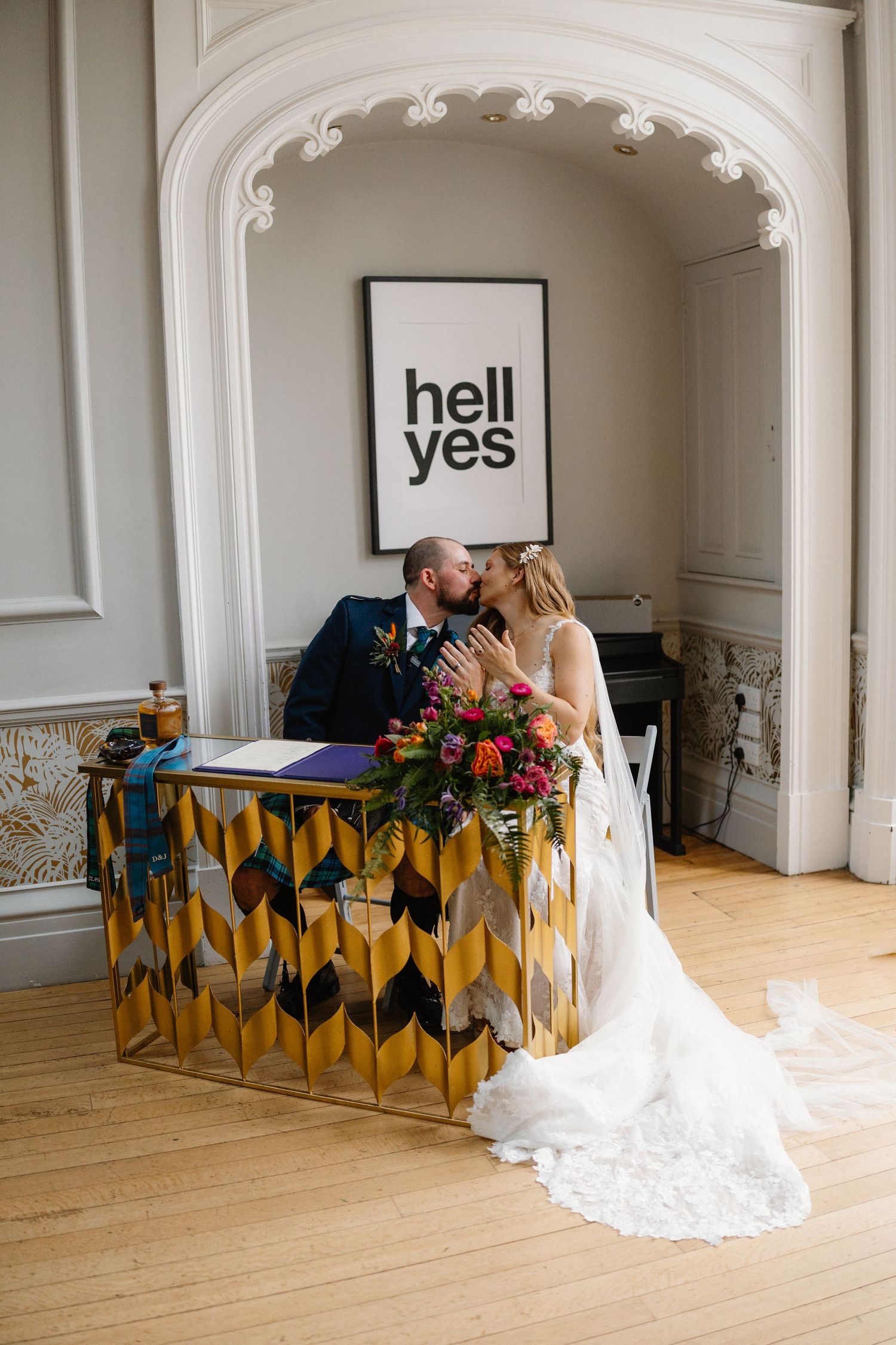Bride and groom share a kiss at a gold geometric desk beneath a bold hell yes sign in an elegant venue.