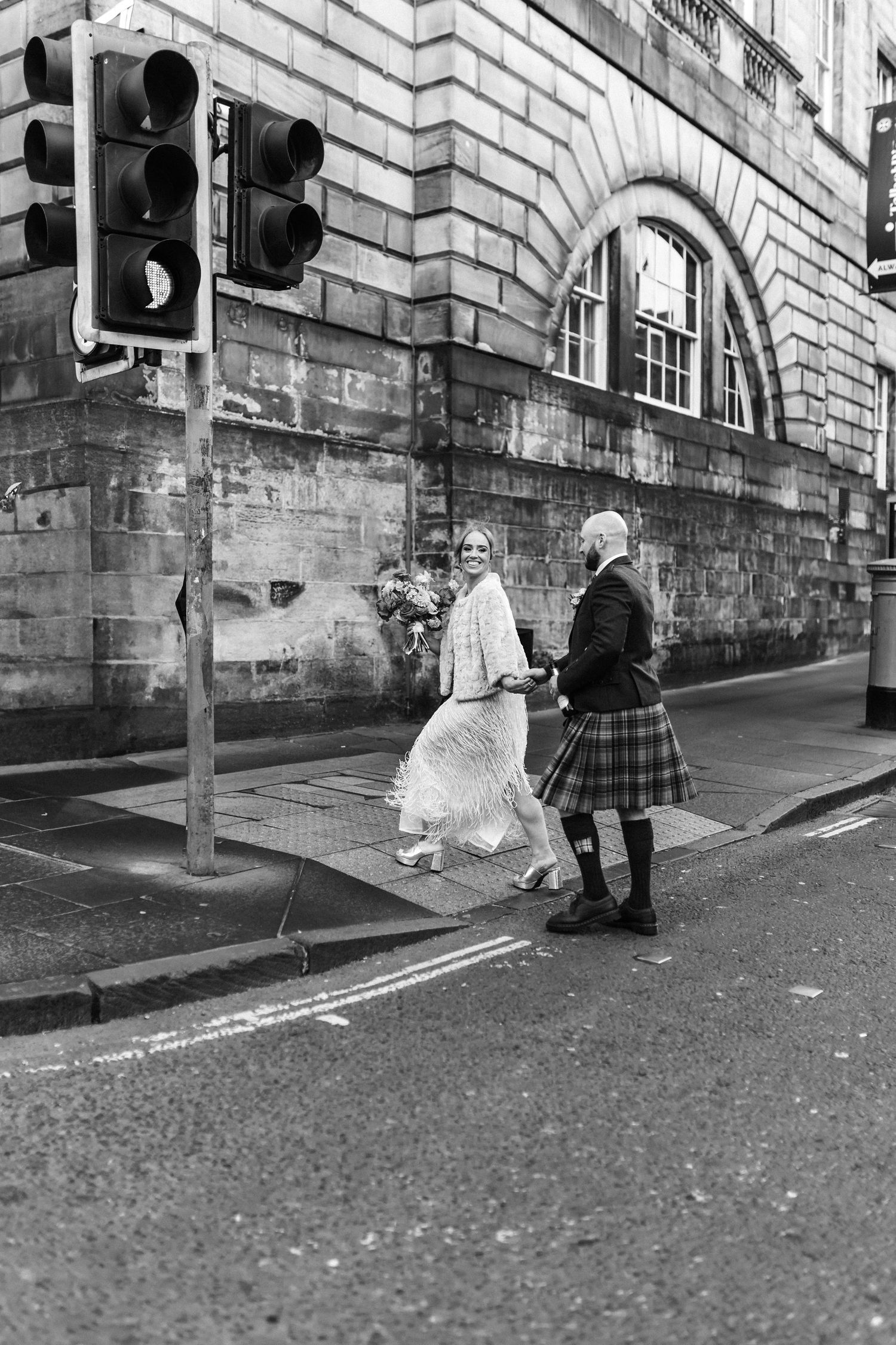 Black and white photo of bride and groom in kilt crossing a cobblestone street in Edinburgh.