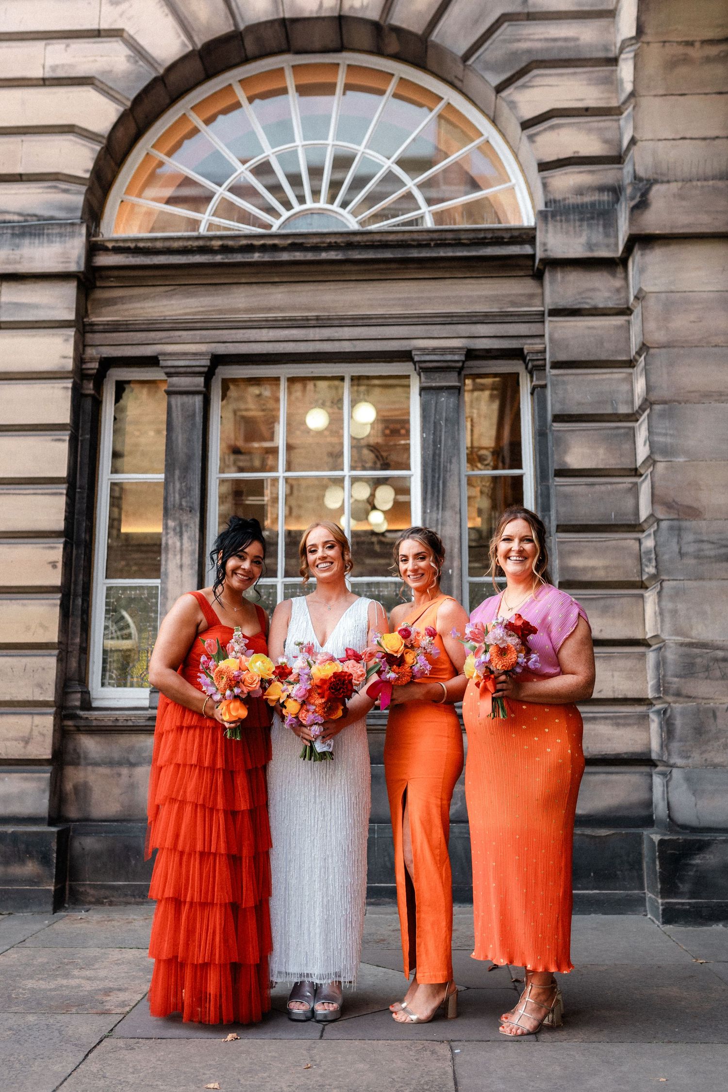 Bride with three bridesmaids in orange dresses holding colorful bouquets outside stone building.