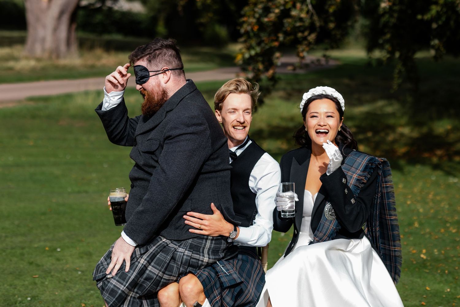 Bride in tartan jacket laughing with groomsmen in kilts at sunny outdoor Scottish wedding.