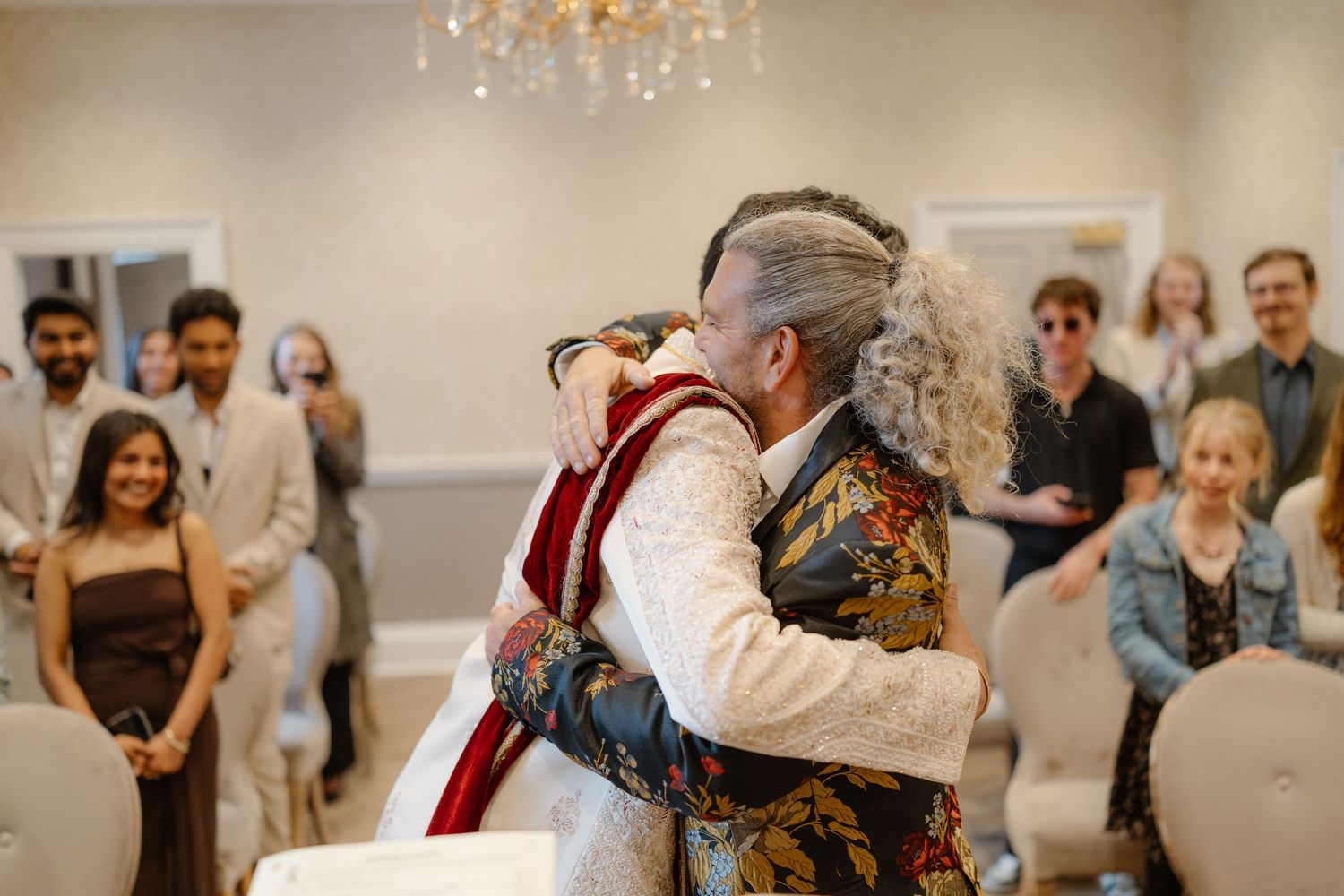 Groom in white sherwani embraces older guest warmly at indoor South Asian wedding reception.