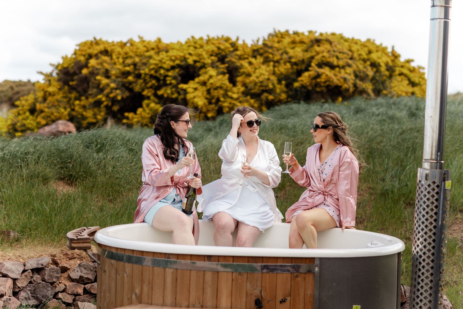Three women in pink robes celebrating with champagne in an outdoor hot tub.