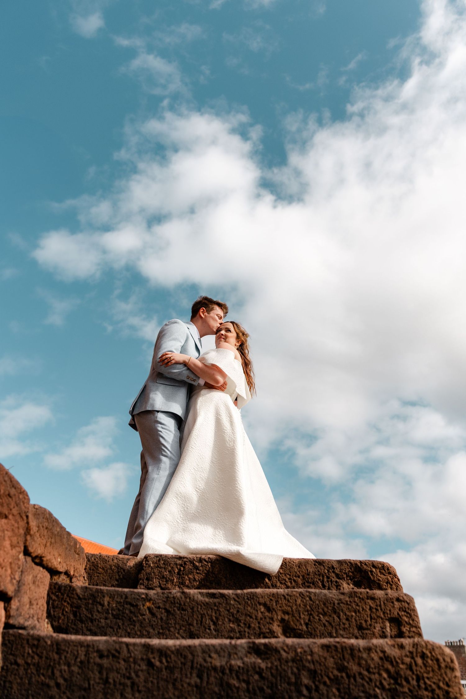 Bride and groom embrace on stone steps against a dramatic blue sky on their wedding day.