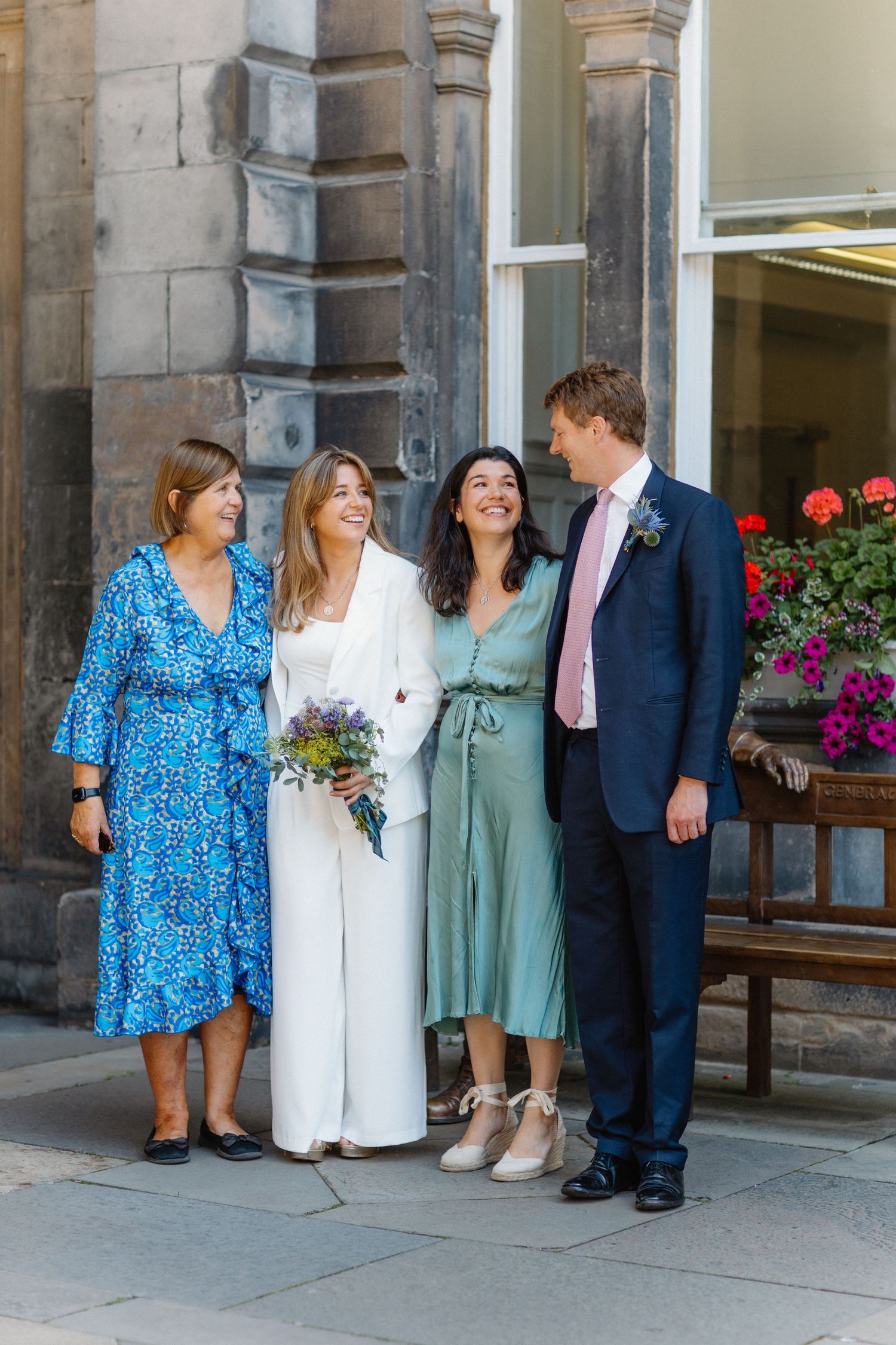 Bride in white pantsuit holding wildflower bouquet posing with family outside a historic stone building.