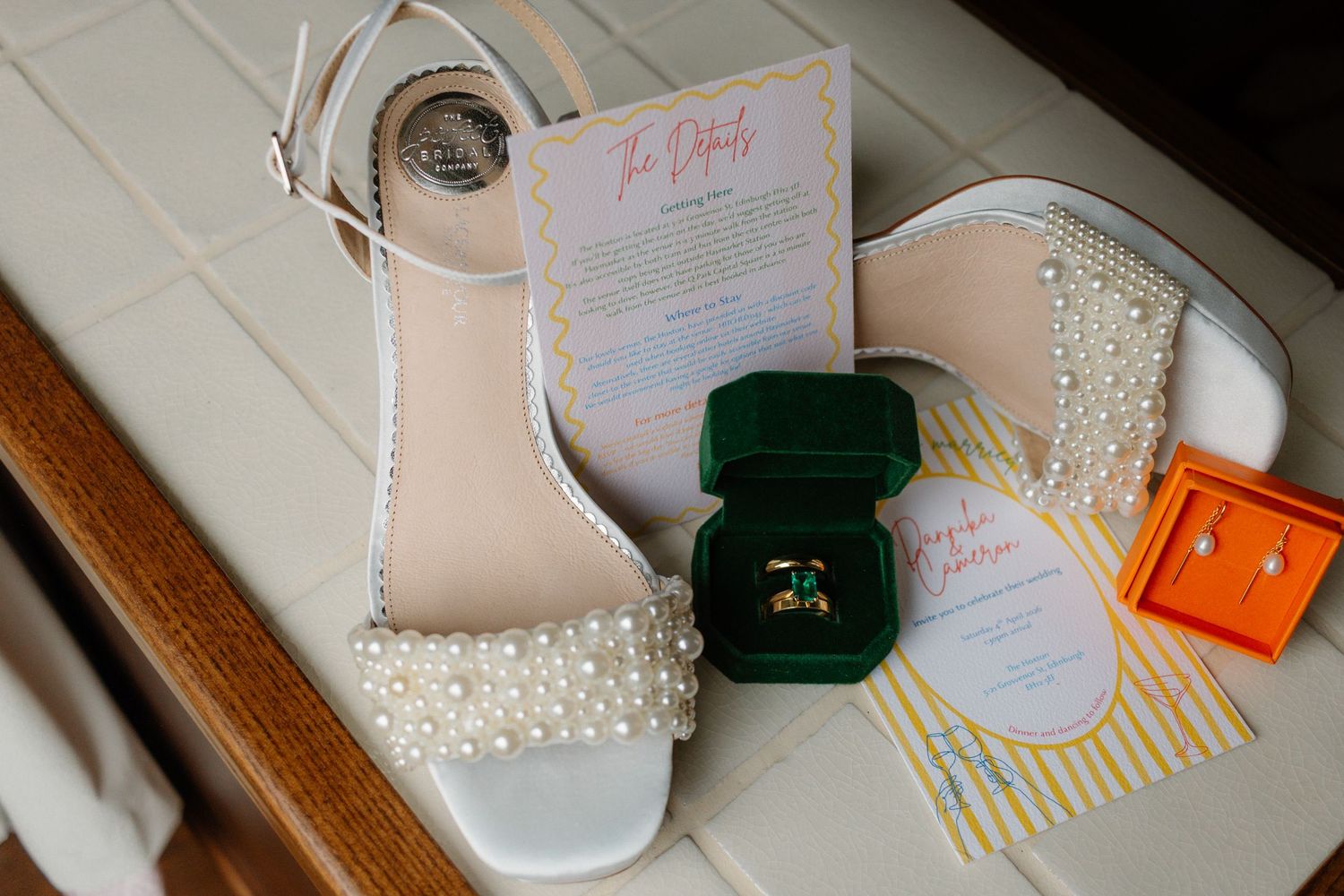 Pearl-adorned white wedding heels displayed with gold rings in a green velvet box, earrings, and colorful wedding stationery.