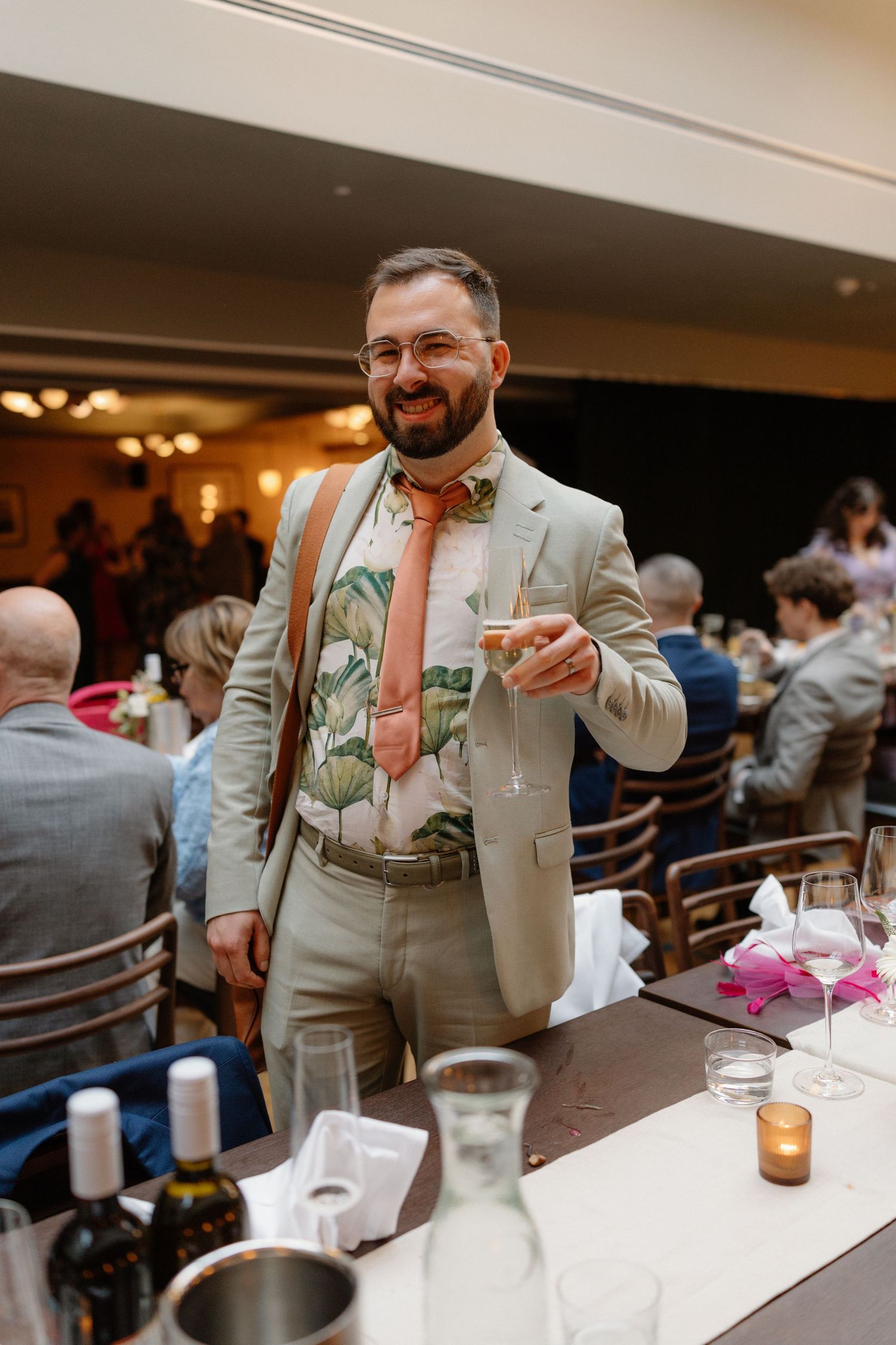 Smiling bearded man in floral shirt and tan suit holding drink at wedding reception.