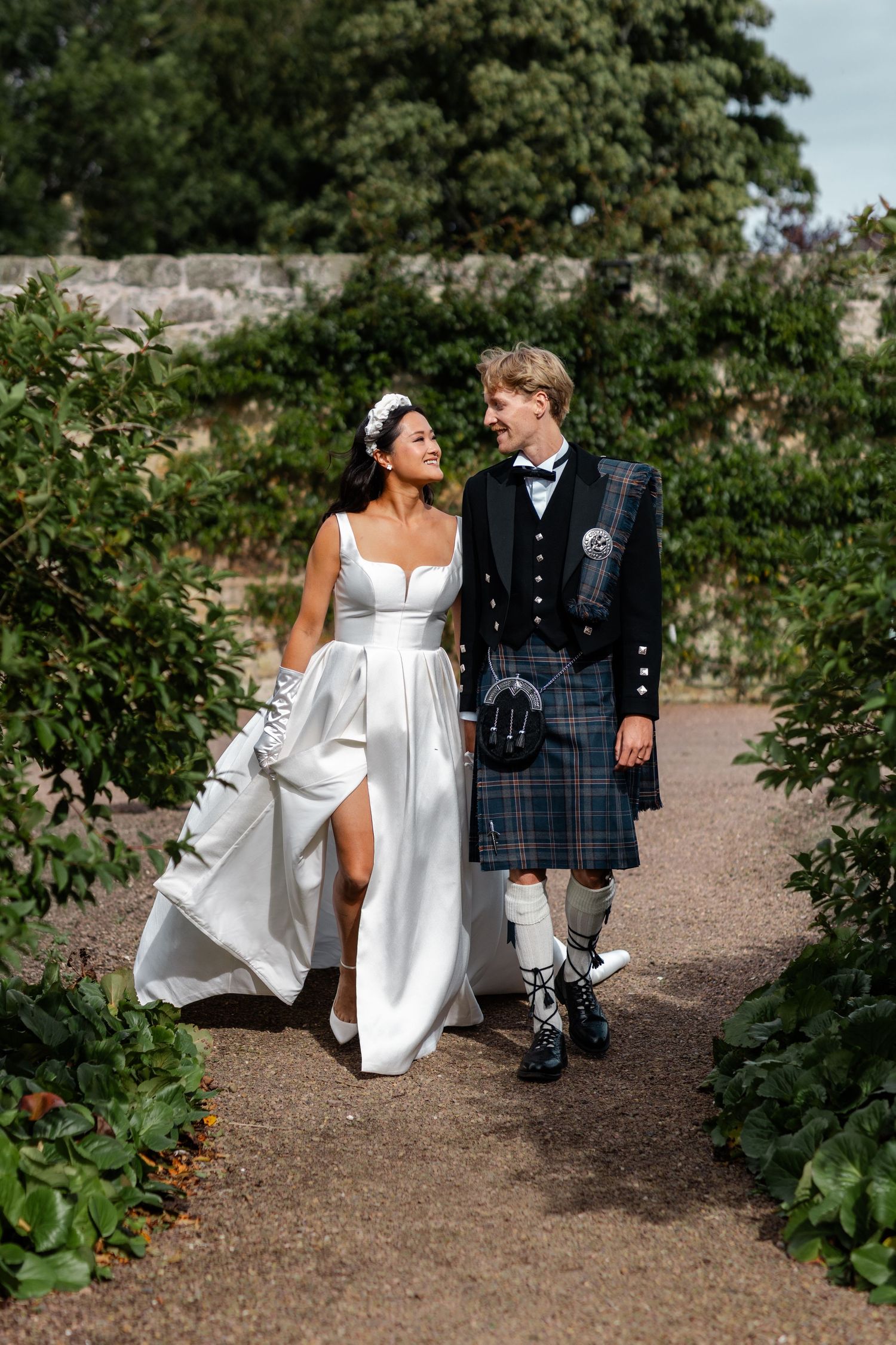Couple walking along garden path wearing traditional Scottish and white wedding attire.