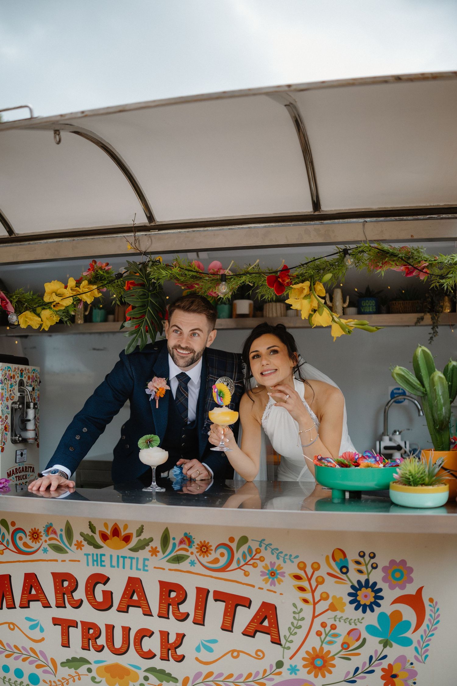 Newlyweds peek through window of colorful margarita food truck decorated with floral designs.