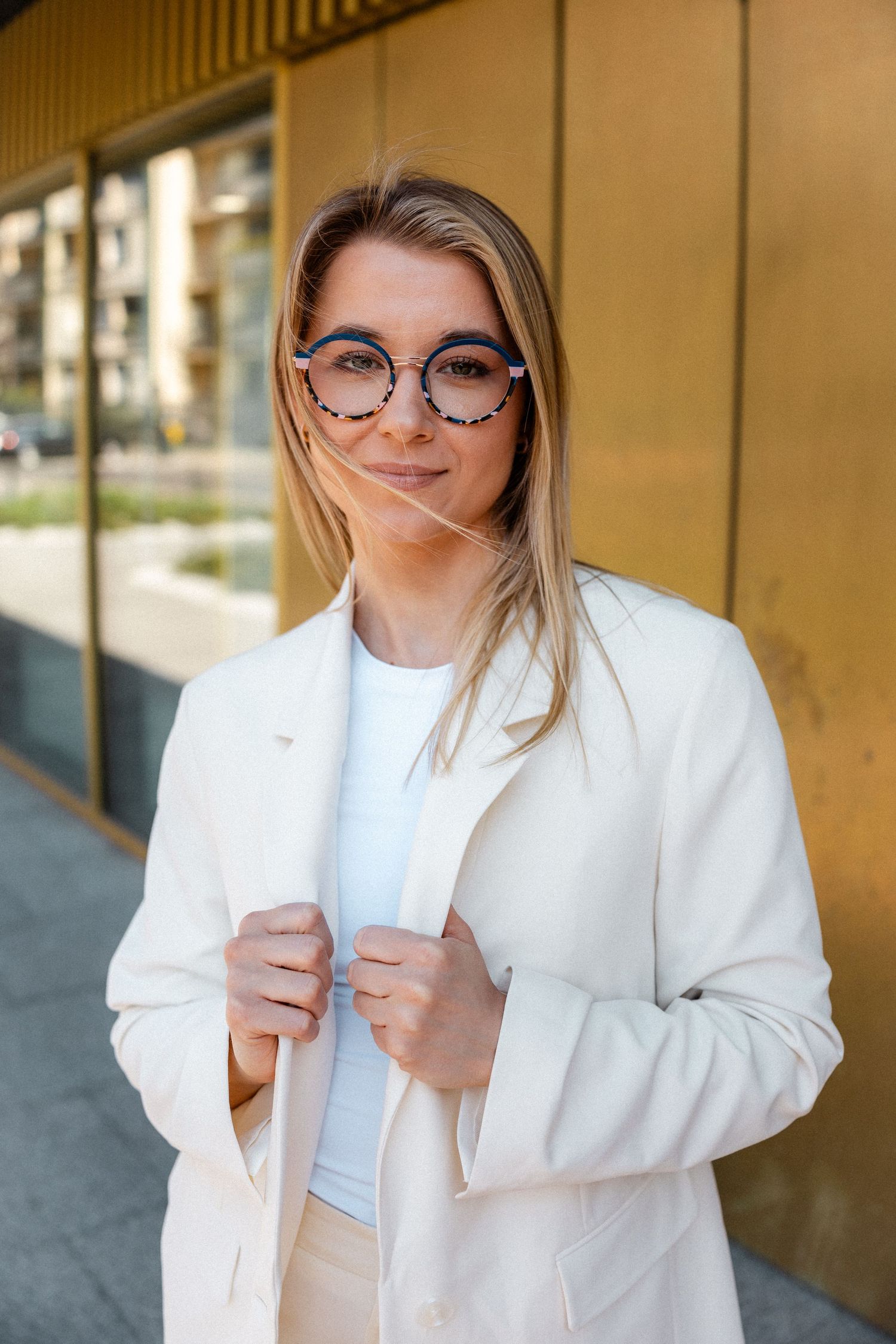 Person in white blazer and glasses posing playfully against metallic wall.
