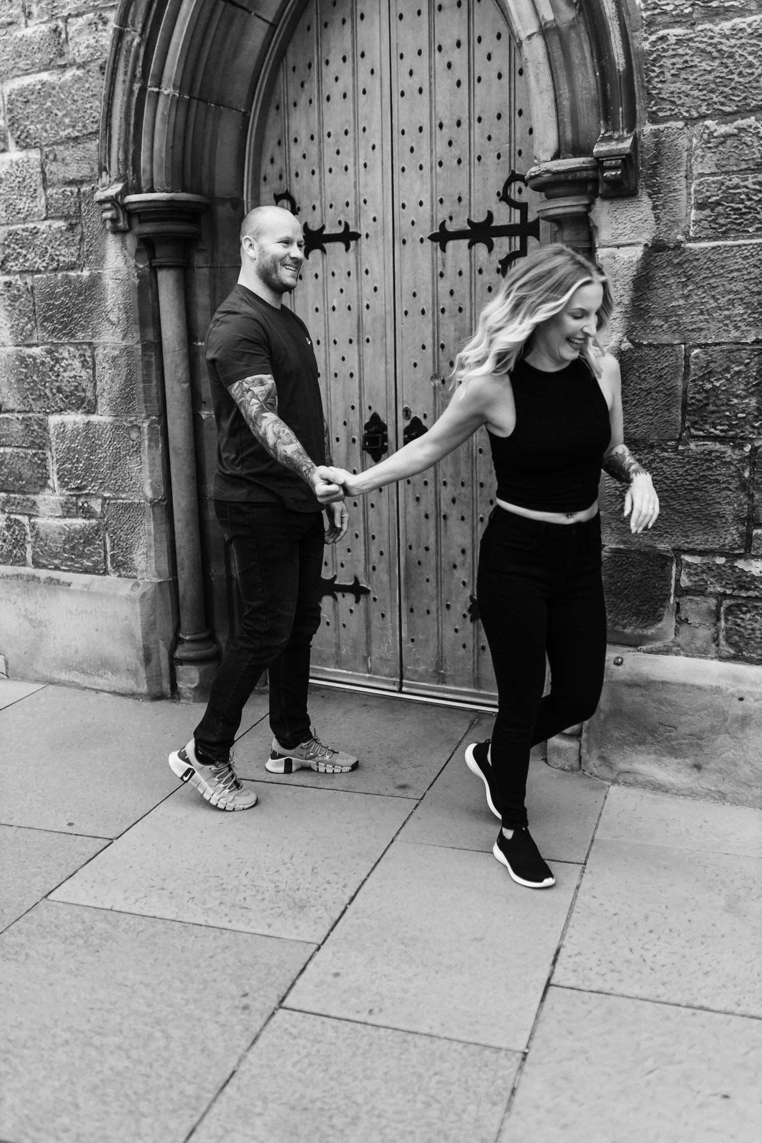 A couple dances and poses playfully in front of a large wooden medieval door.