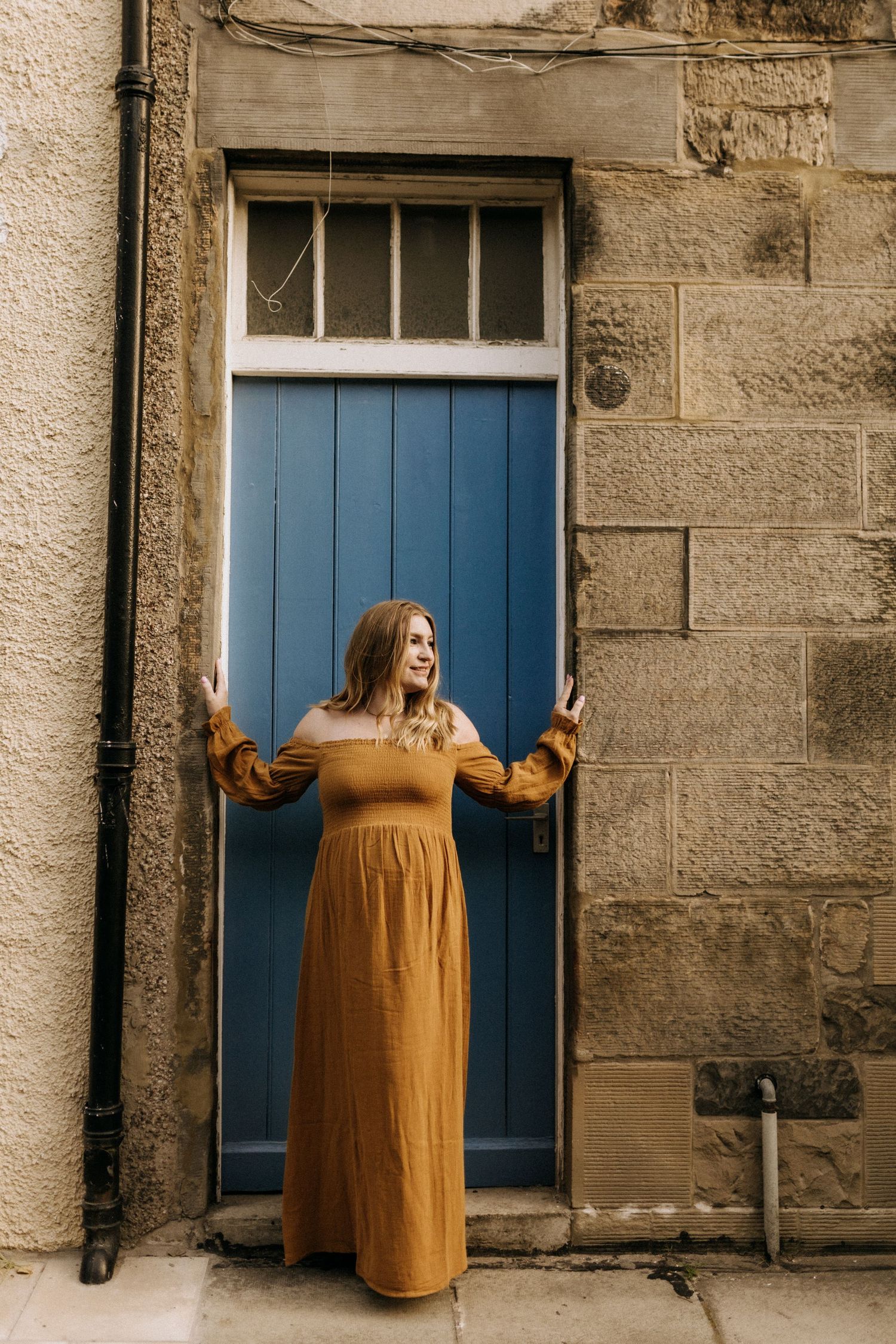 A person in a mustard dress poses against stone walls with blue doors.