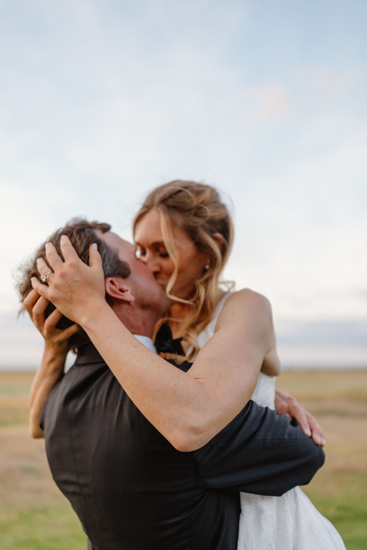 Bride and groom sharing passionate kiss outdoors under soft pastel evening sky at golden hour.